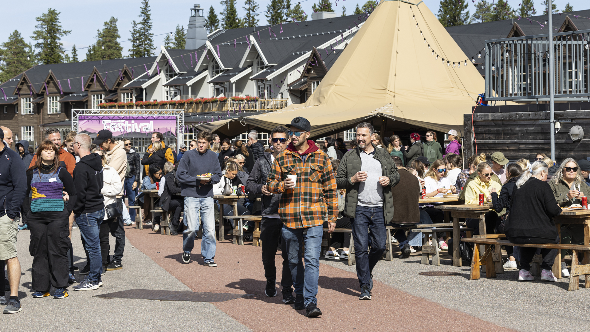 Festivalbesøkende nyter solfylt dag på utendørs kick off med boder og servering, omgitt av tradisjonelle norske bygninger og trær i bakgrunnen