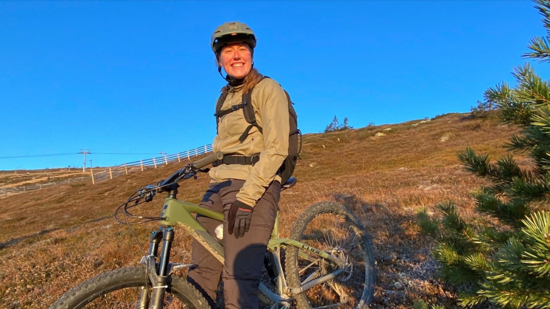En person på mountainbike på en solrig dag i naturen, iført en brun jakke og hjelm, på en skråning med tørret græs og blå himmel i baggrunden