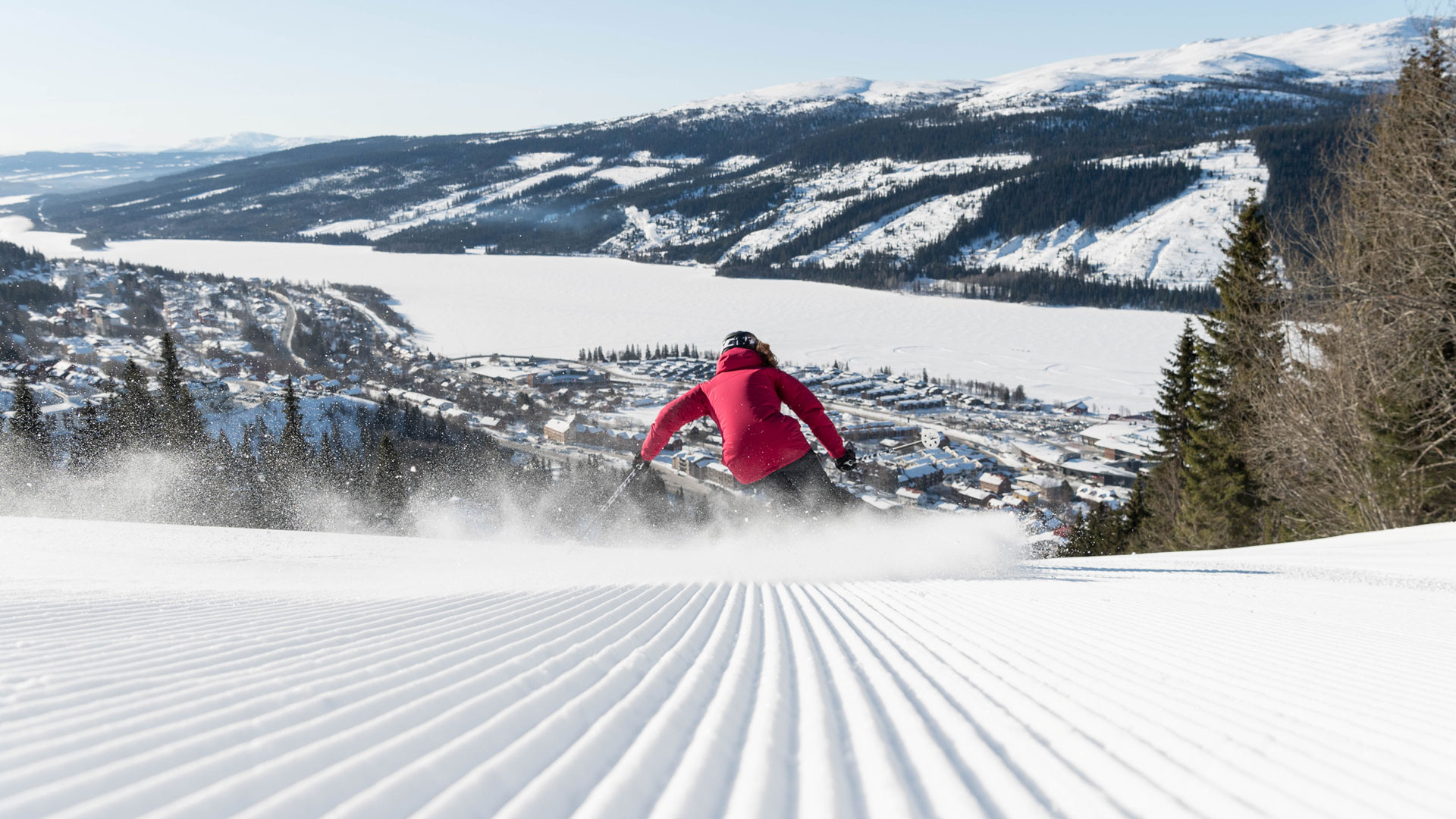 Skiløber i rød jakke står på ski ned ad en snedækket bakke med udsigt over landsbyen Åre og de omkringliggende fjelde