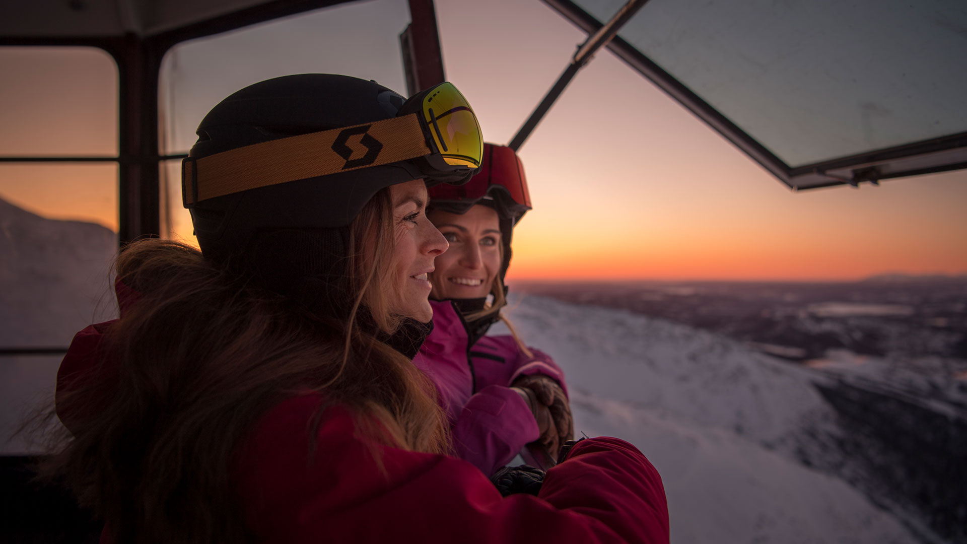 To personer i skitøj ser ud fra en skilift mod solnedgangen over snedækkede bjerge