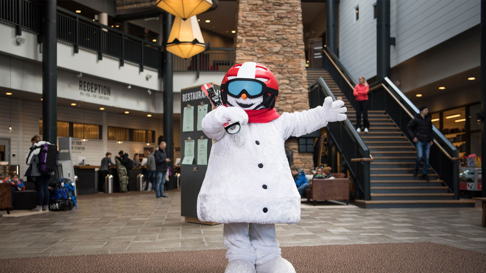 Maskot i snedragt poserer i en hotellobby med reception og restaurant i baggrunden