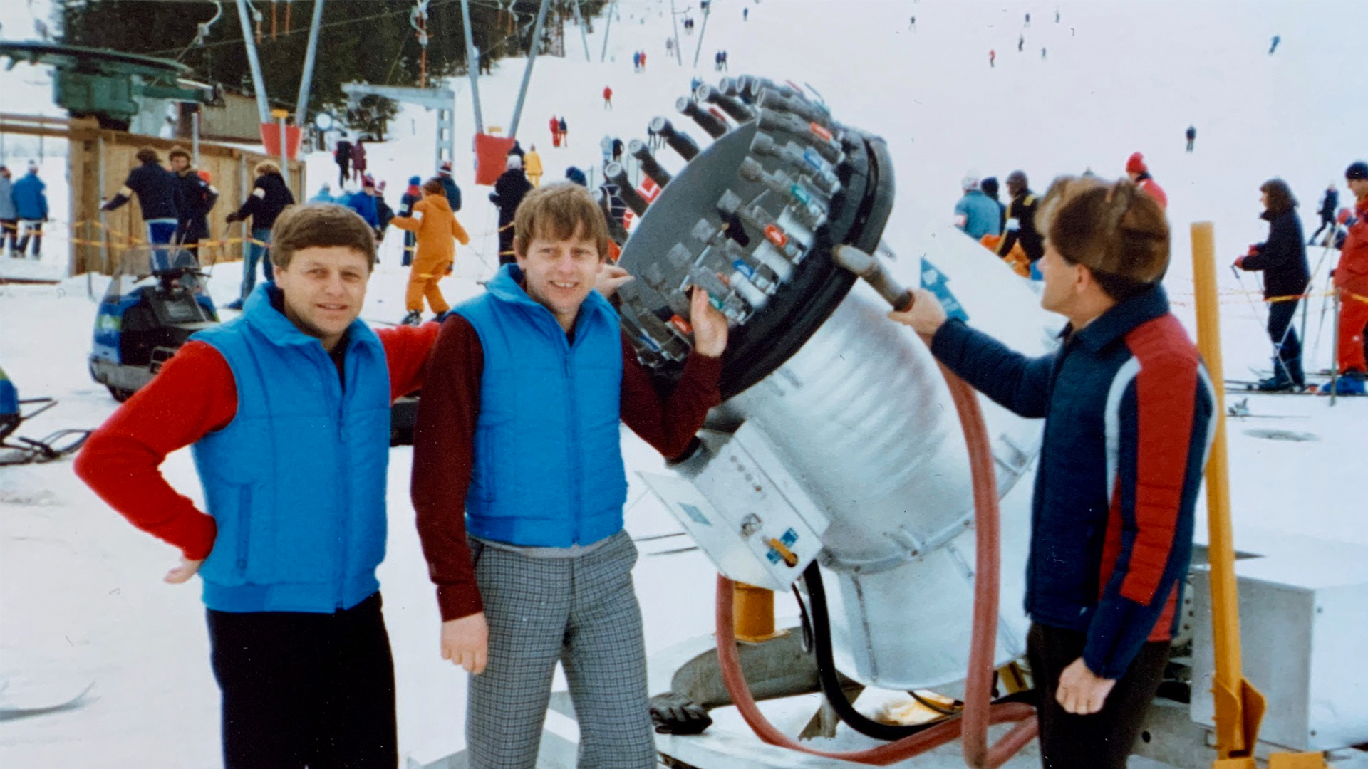 Tre personer i blå västar vid en snökanon på en skidort. Bakom dem ser man skidåkare och en lift