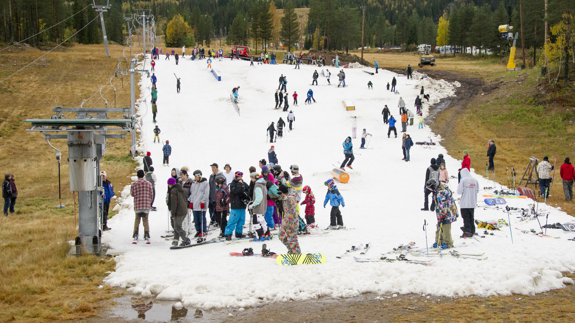 Skiers gathered on a snowy slope in a ski resort, surrounded by autumn-colored trees and grass