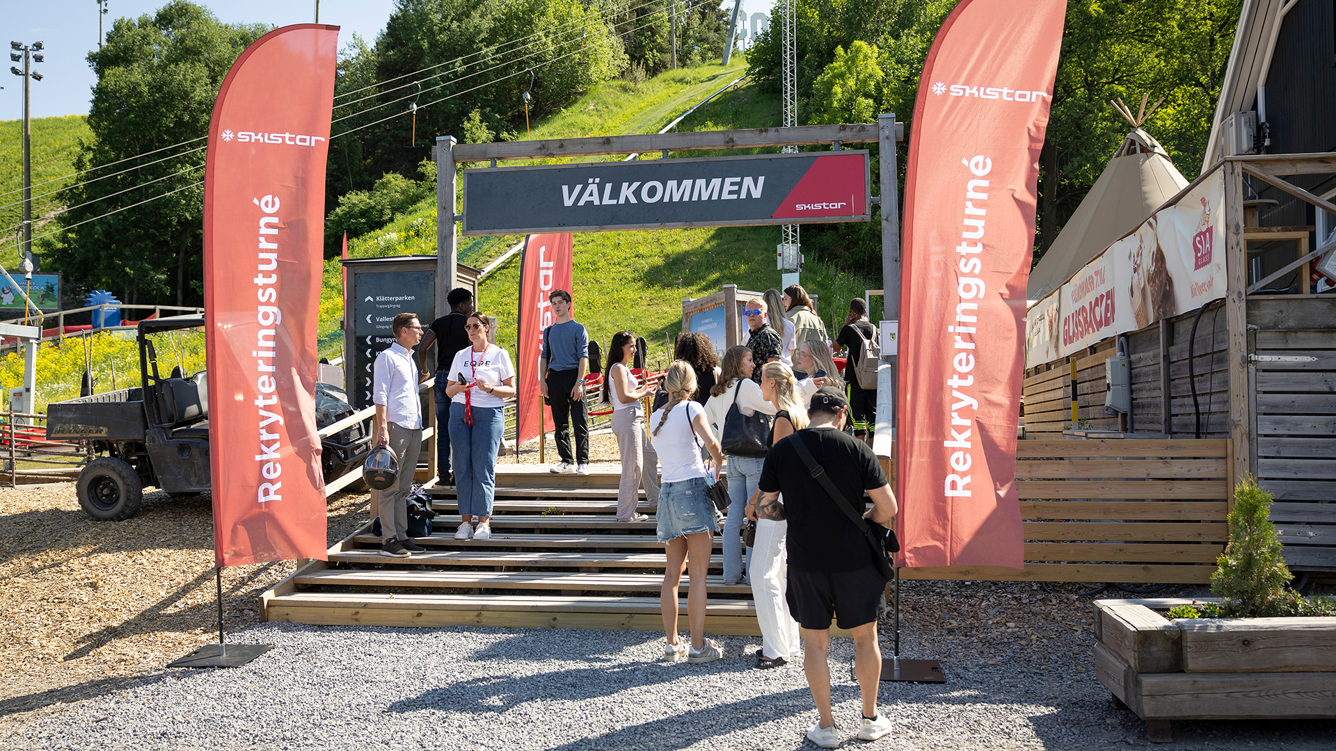 Besökare samlas vid Skistars rekryteringsturné under en solig dag, med välkomstskylt och röda flaggor vid en utomhuspark