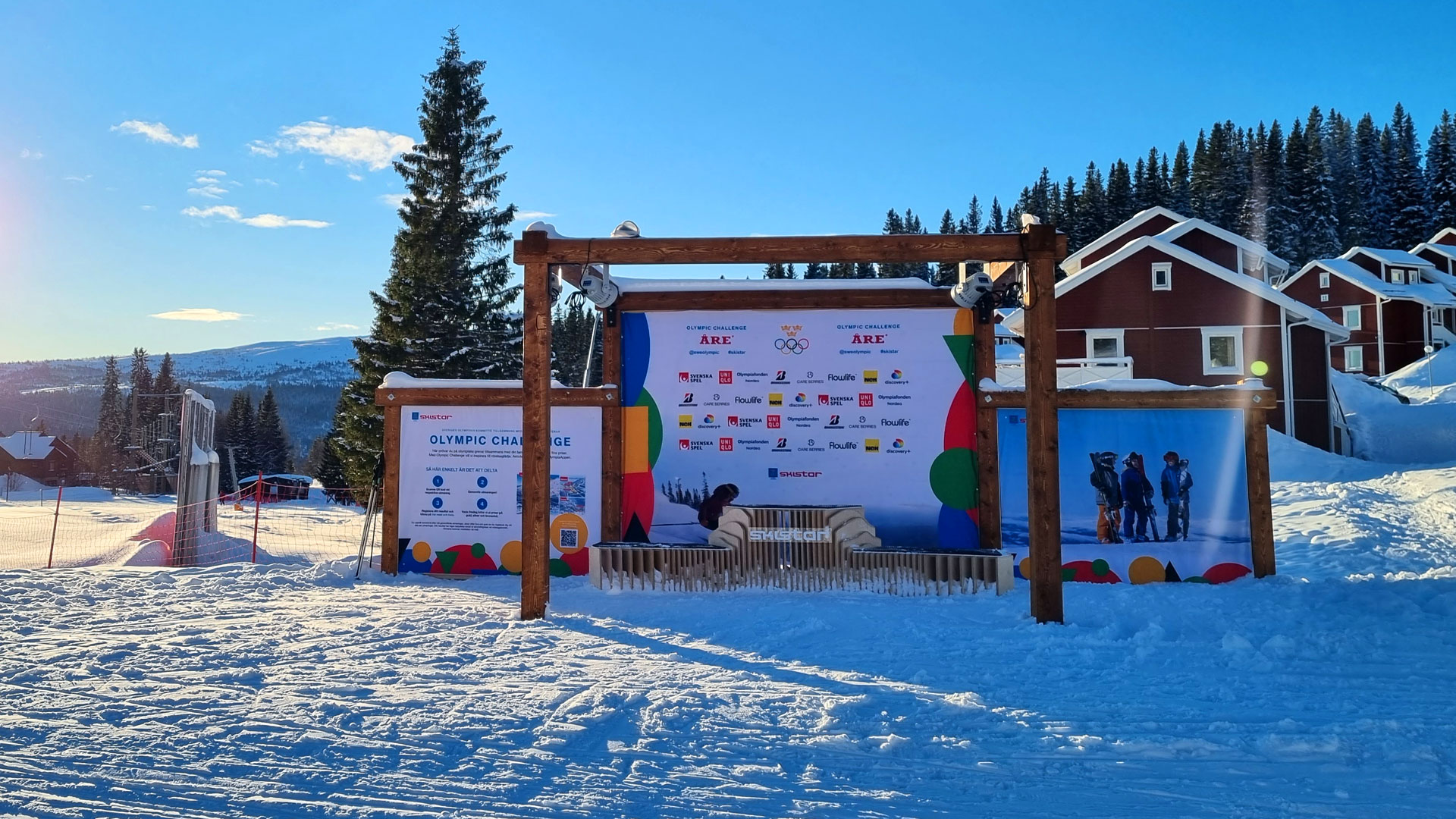 Snötäckt landskap med skidtävling i Åre, Sverige. Stor reklamvägg med sponsorer i bakgrunden, omgiven av vinterstugor och skog under en klarblå himmel