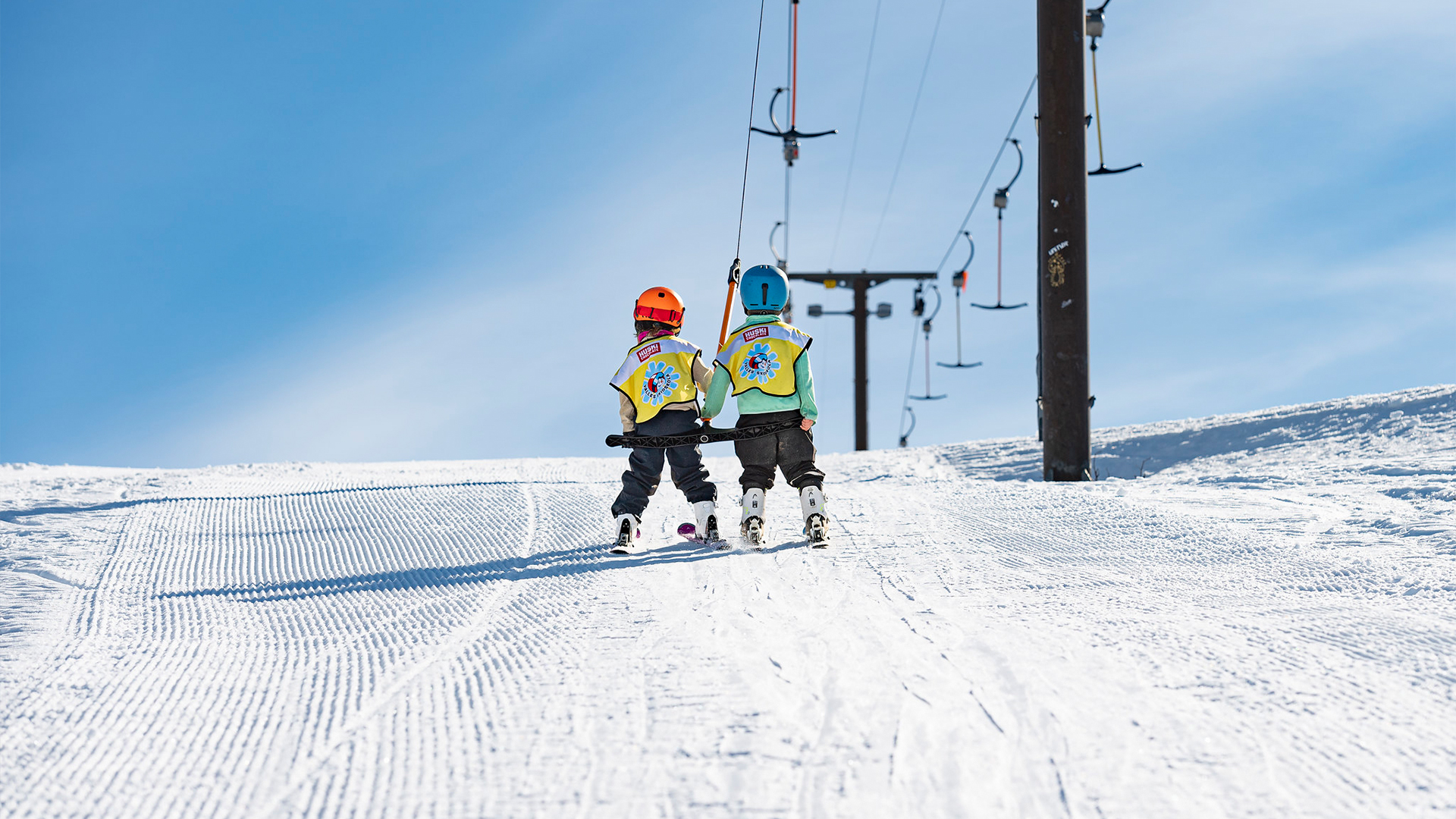 Two children in colorful ski gear ride a ski lift up a snowy slope under a clear blue sky