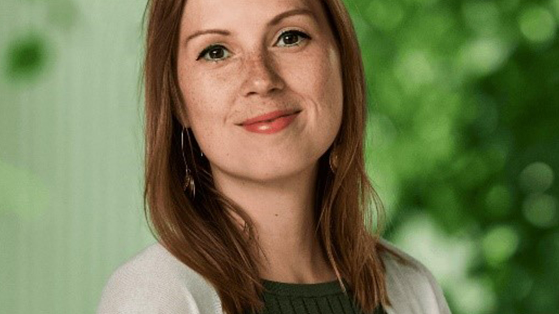Kvinna med brunt hår och grön tröja ler mot kameran i en grönskande bakgrund