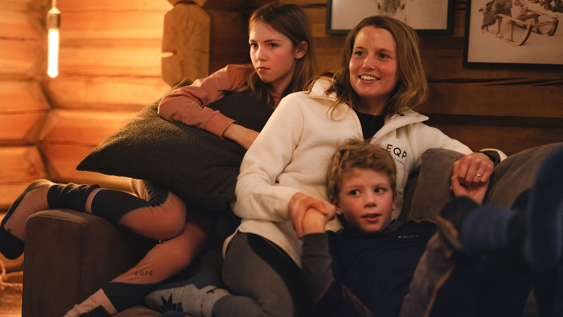 Familj myser i en trästuga, klädda i vinterkläder, sittandes på en soffa med varm belysning i bakgrunden