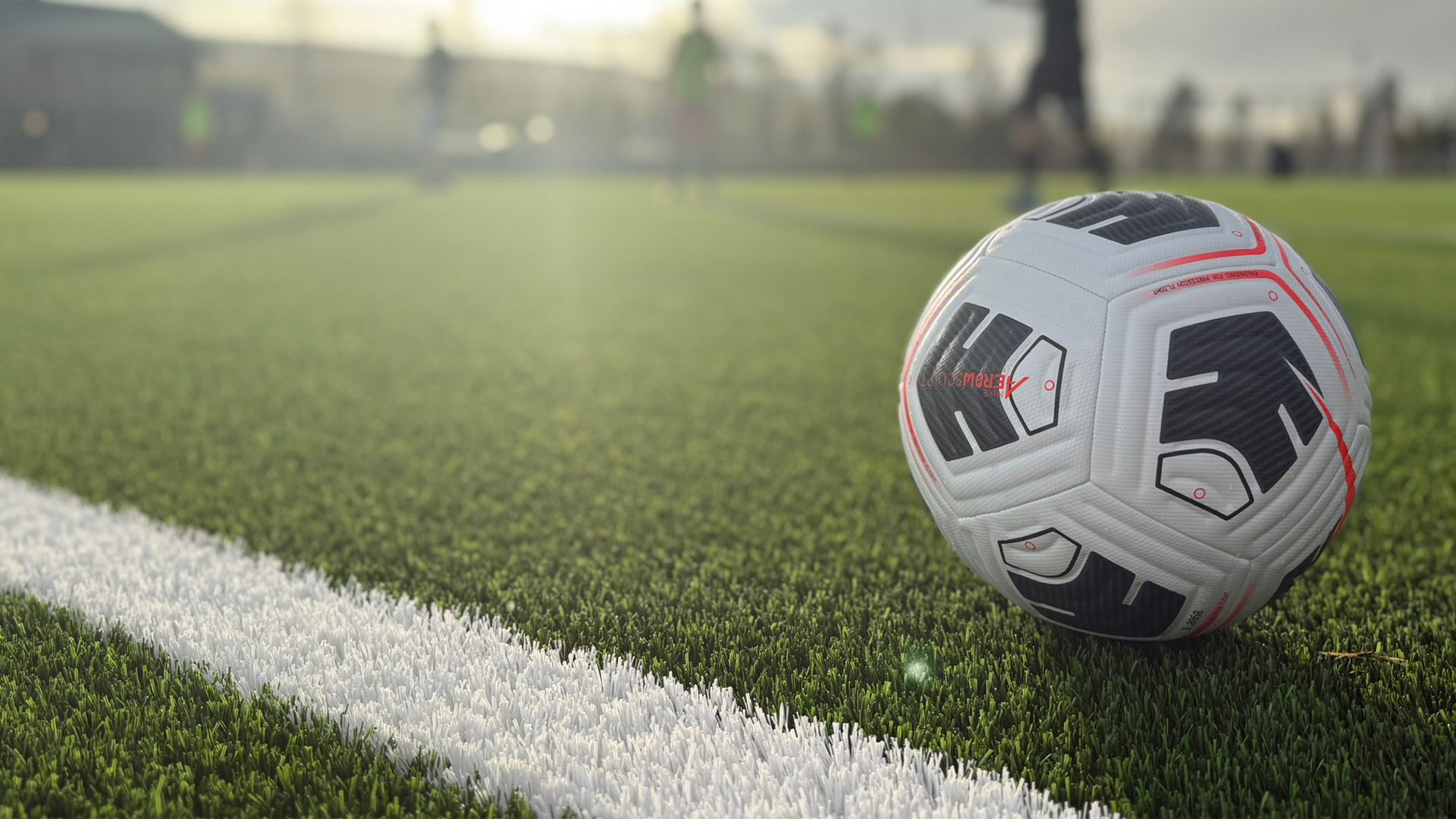 Fodbold på grøn kunstgræsbane tæt på hvid linje, sollys i baggrunden