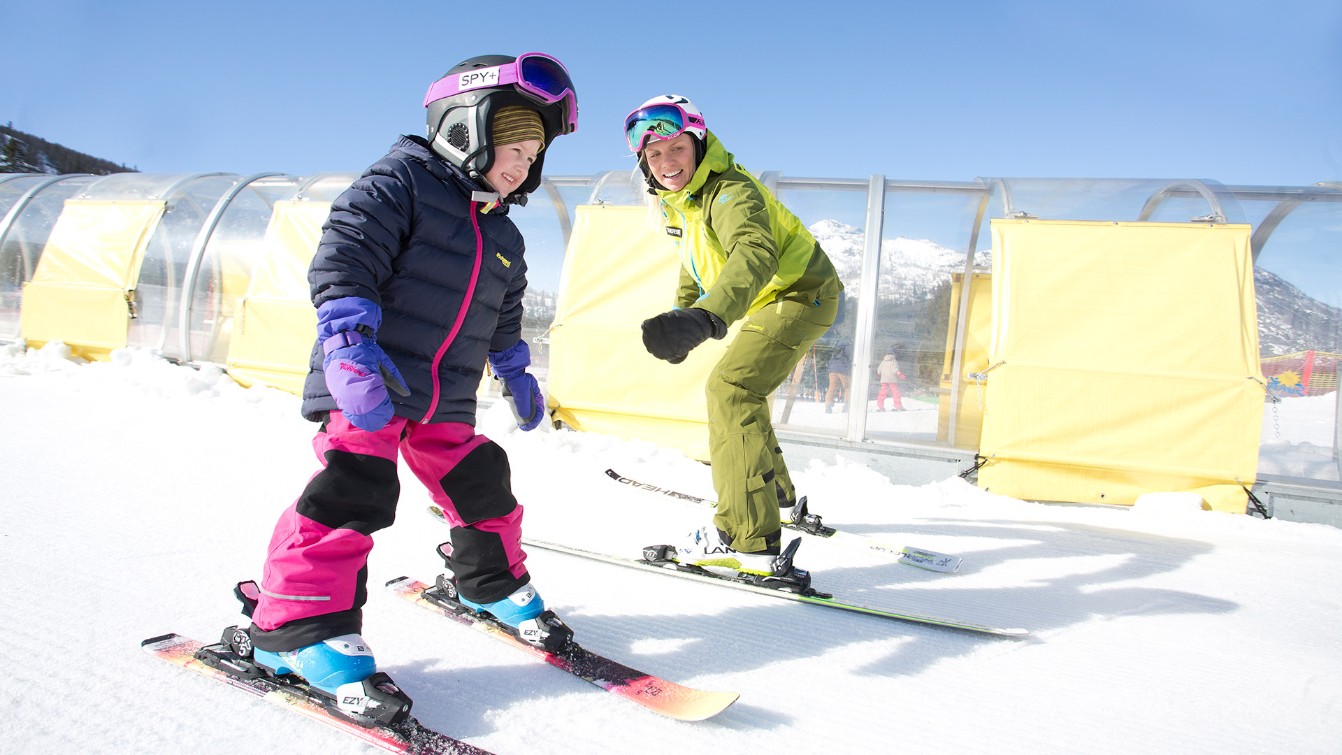 Barn och vuxen åker skidor i soligt väder på en skidort, med tunnelbana och snöklädda berg i bakgrunden