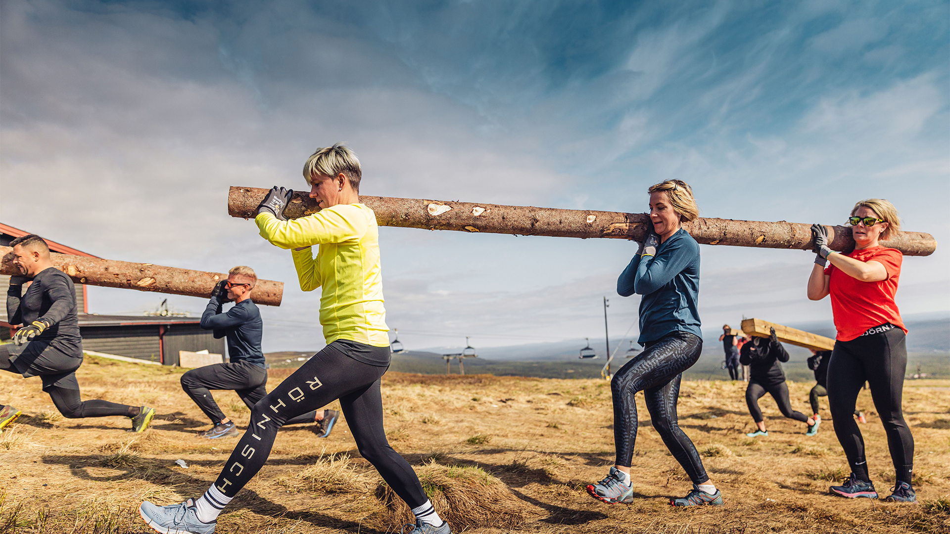Mennesker deltar i en utendørstreningsøvelse og bærer stokker sammen på en solfylt dag i fjellet