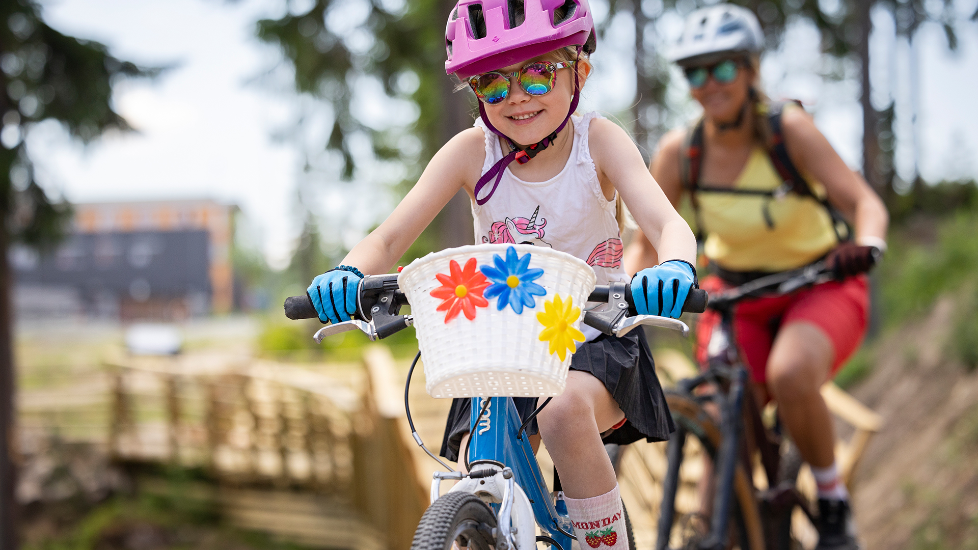 Flicka med rosa hjälm och cykelkorg dekorerad med blommor cyklar med vuxen på stig i skogen, 