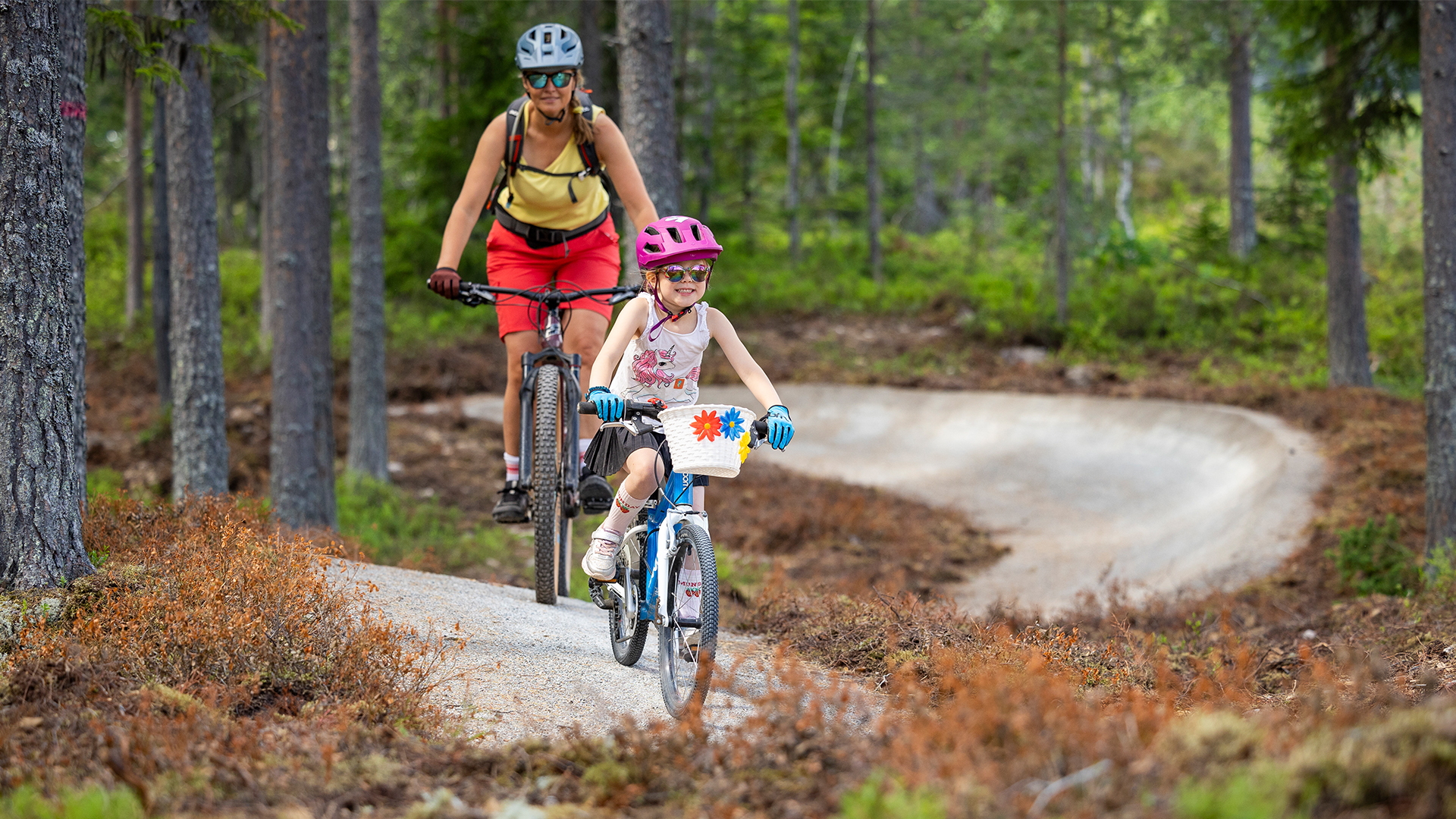 Barn och vuxen cyklar på en cykelled i skogen, båda bär cykelhjälmar och ser glada ut