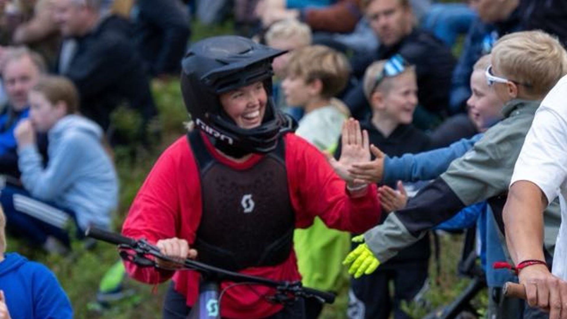 Cykelrytter i rød trøje og hjelm giver high-five til tilskuere under cykelløb. Publikummet er begejstret og engageret