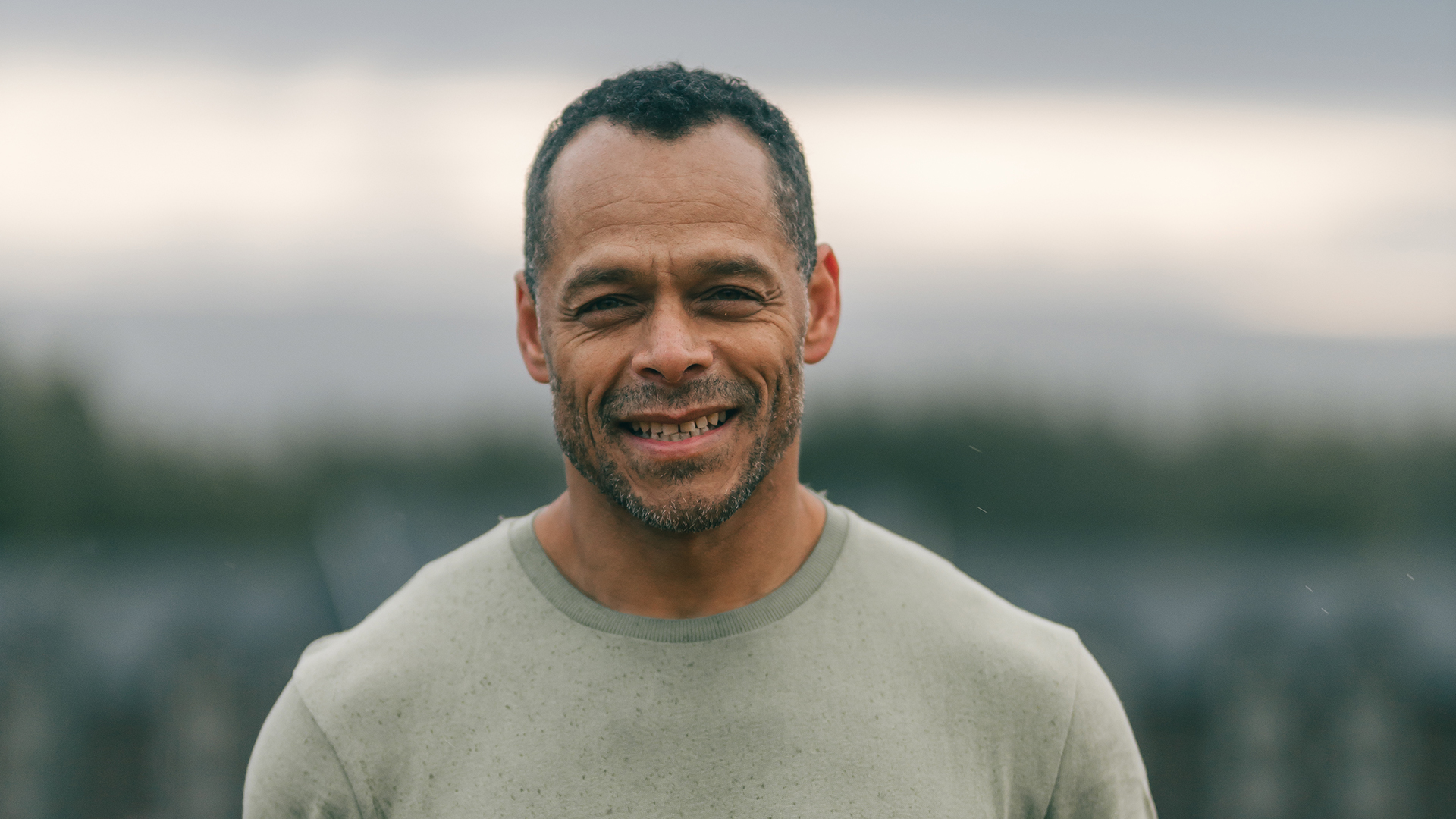 Leende man med kort hår och skäggstubb, iklädd en ljusgrön t-shirt utomhus med suddig bakgrund