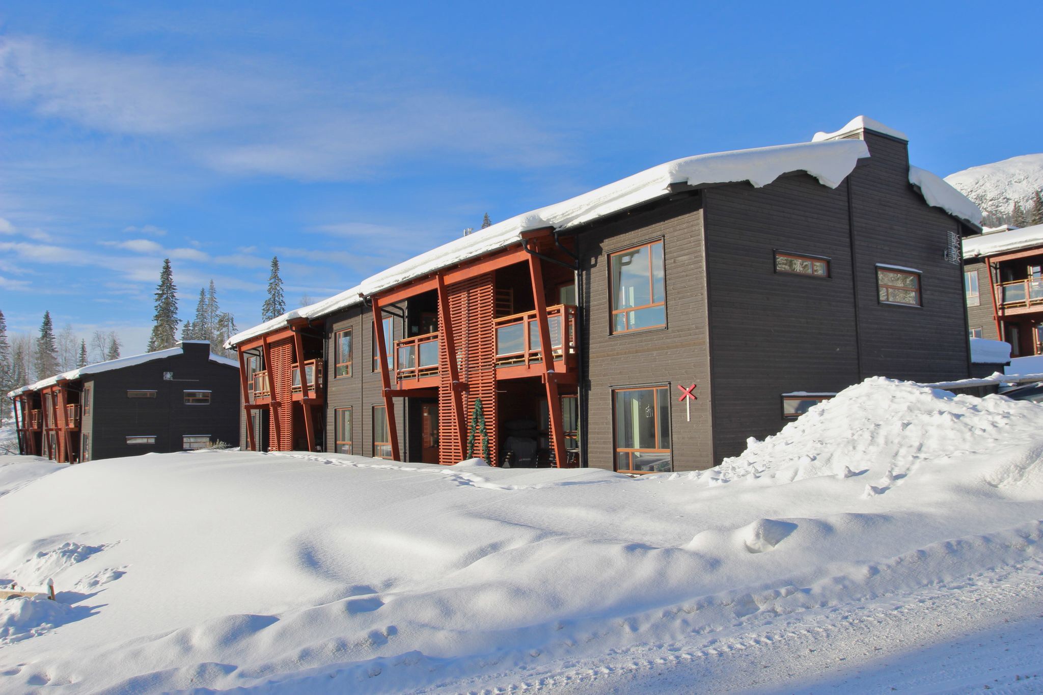 Modern fjällstuga täckt av snö i fjällen med klarblå himmel och granar i bakgrunden