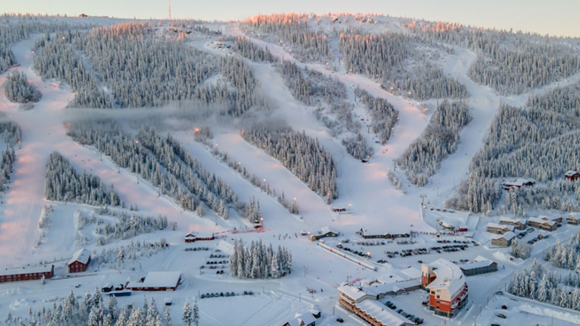 Flygfoto över snöklädda skidbackar och skog i ett vinterlandskap med byggnader vid foten av berget, troligen en skidanläggning i Sverige