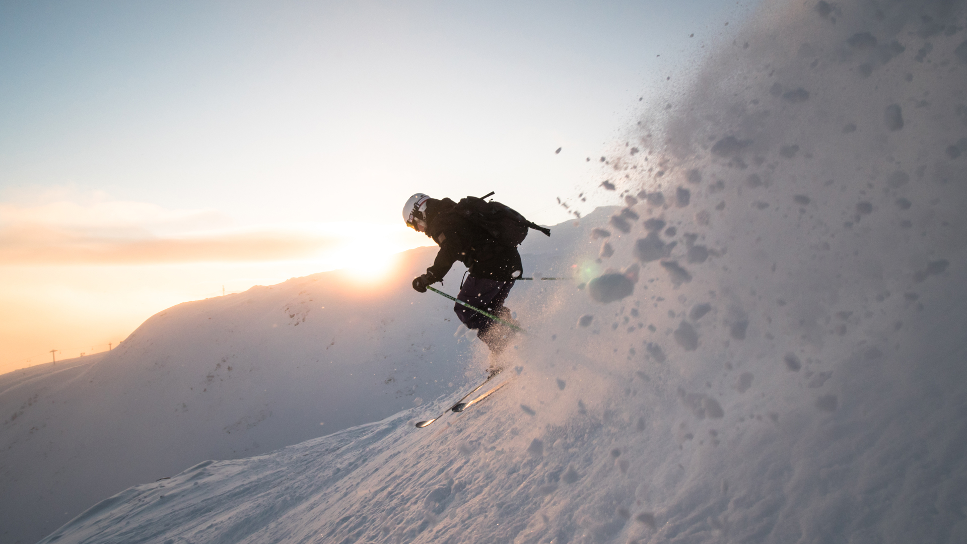 Person åker skidor nerför en snötäckt bergssida i soluppgång, skapar snömoln