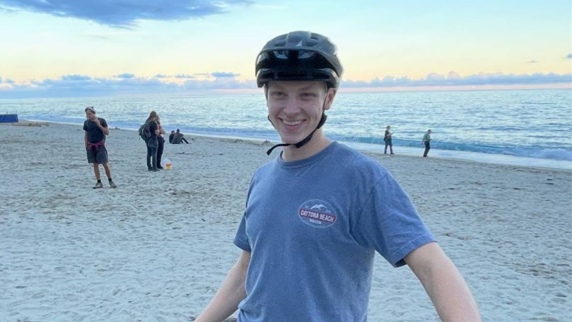 Ung mand med hjelm smiler på stranden ved solnedgang, med havet i baggrunden og folk der går langs kystlinjen