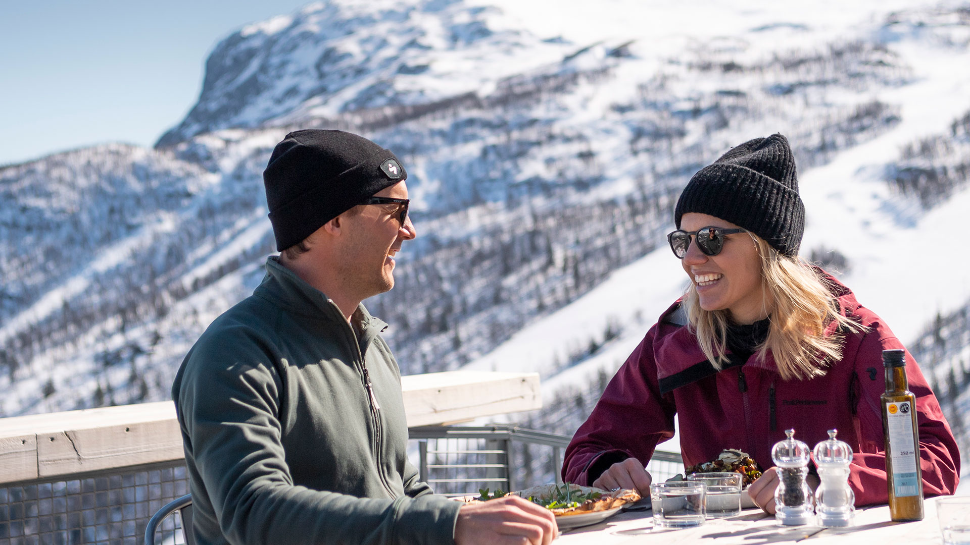 Två personer njuter av en måltid utomhus med en snötäckt bergsutsikt i bakgrunden