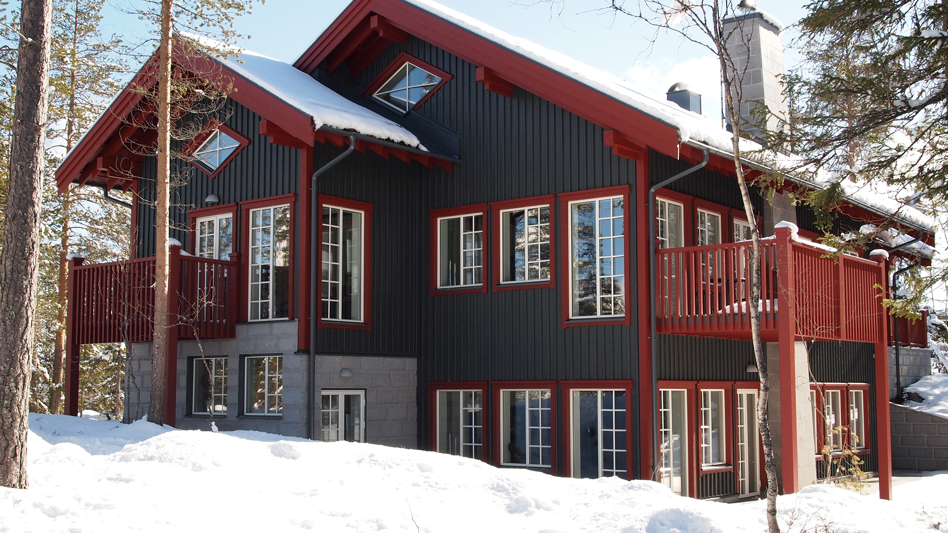 Fjällstuga i trä med snötäckta tak och balkong, omgiven av tallar och snö, i vinterlandskap i Sverige