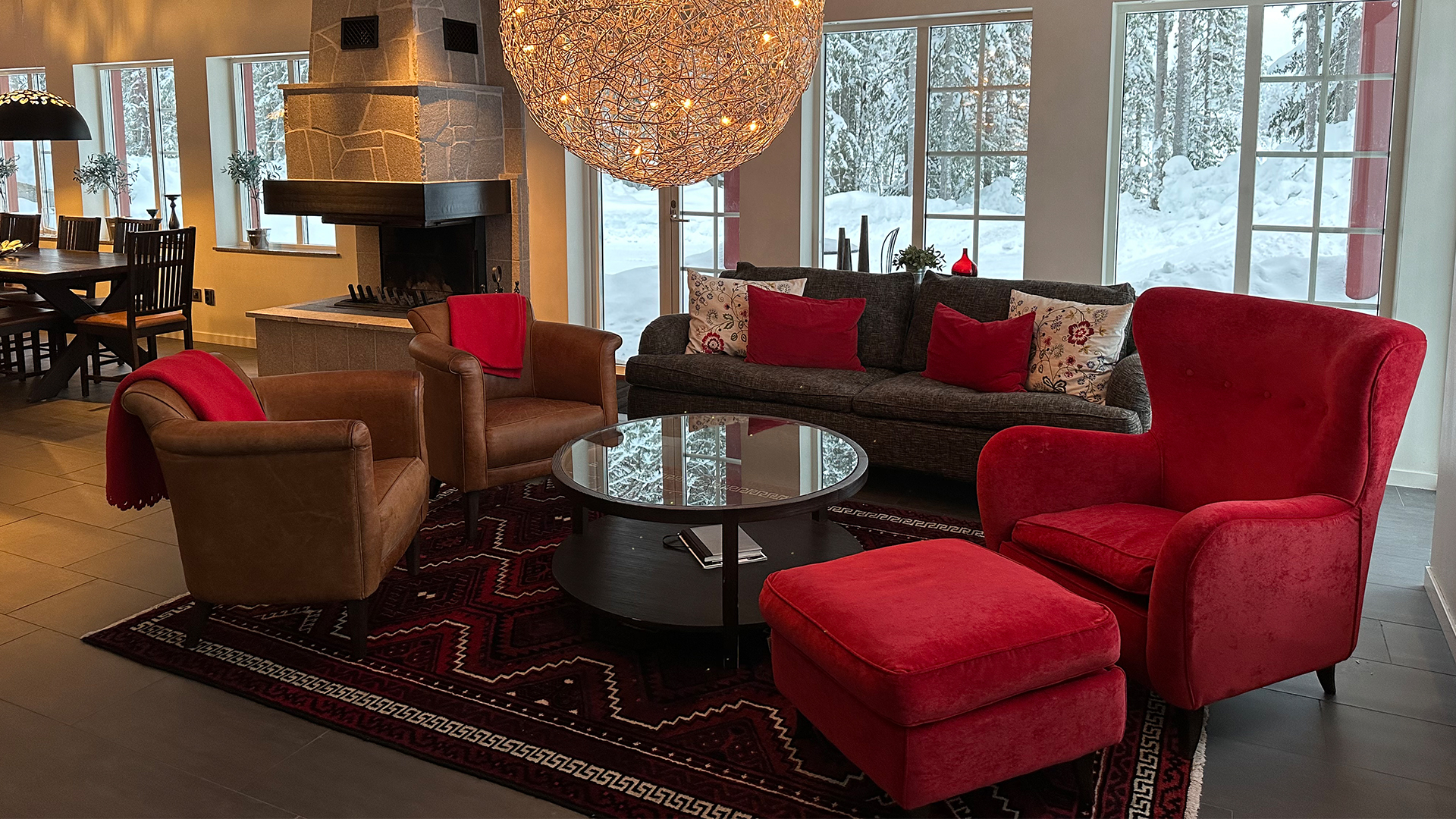 Vardagsrum med röda fåtöljer, soffa med prydnadskuddar, glasbord, och stor taklampa. Eldstad och stora fönster med snöutsikt