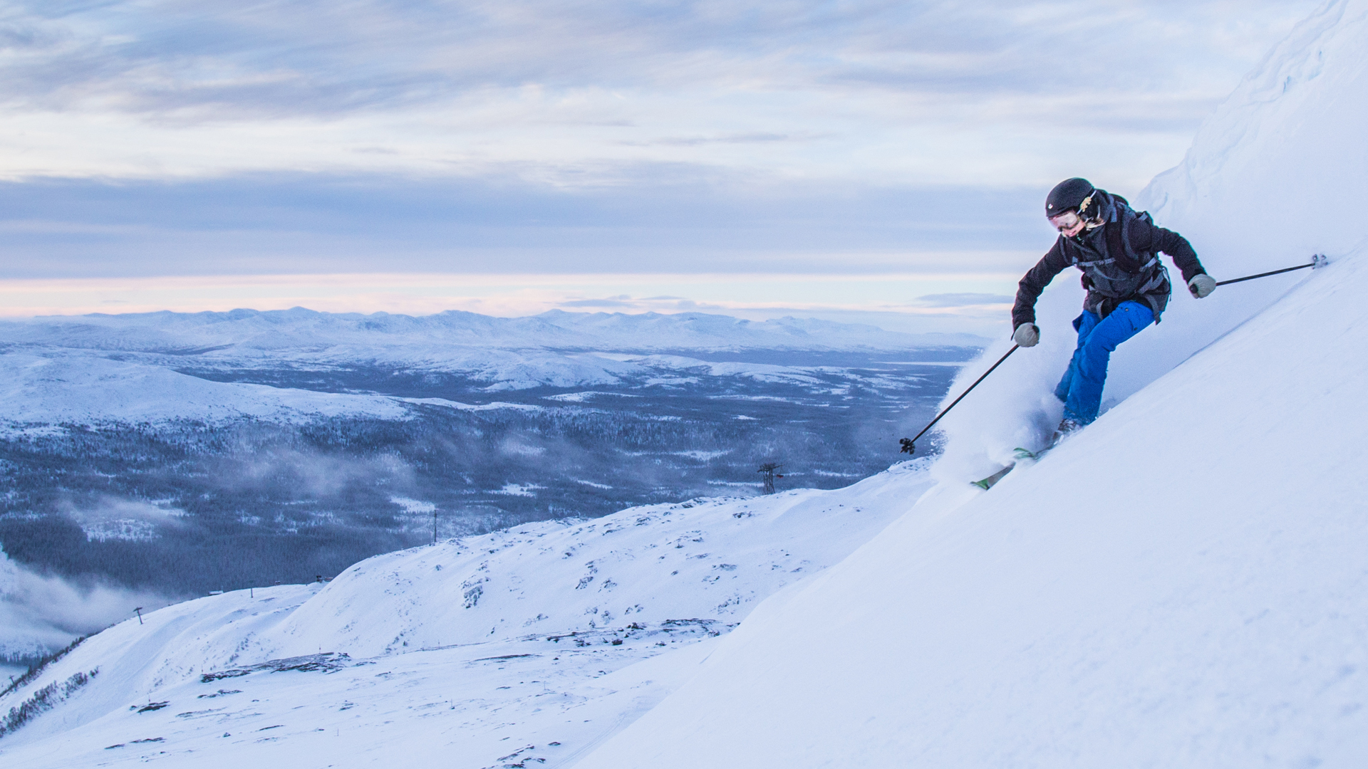 Person åker skidor nedför en brant, snöklädd backe med en vidsträckt utsikt över bergslandskap i bakgrunden, under en molnig himmel