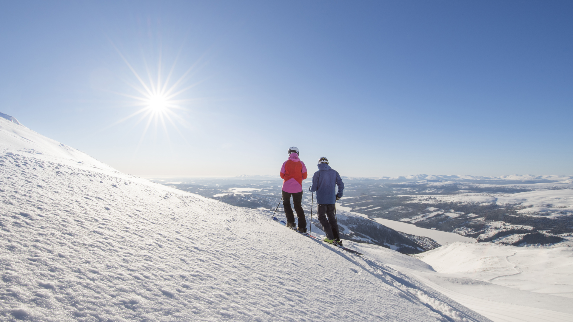 Två skidåkare på en solig bergssluttning med snö och vy över ett vinterlandskap i fjällen