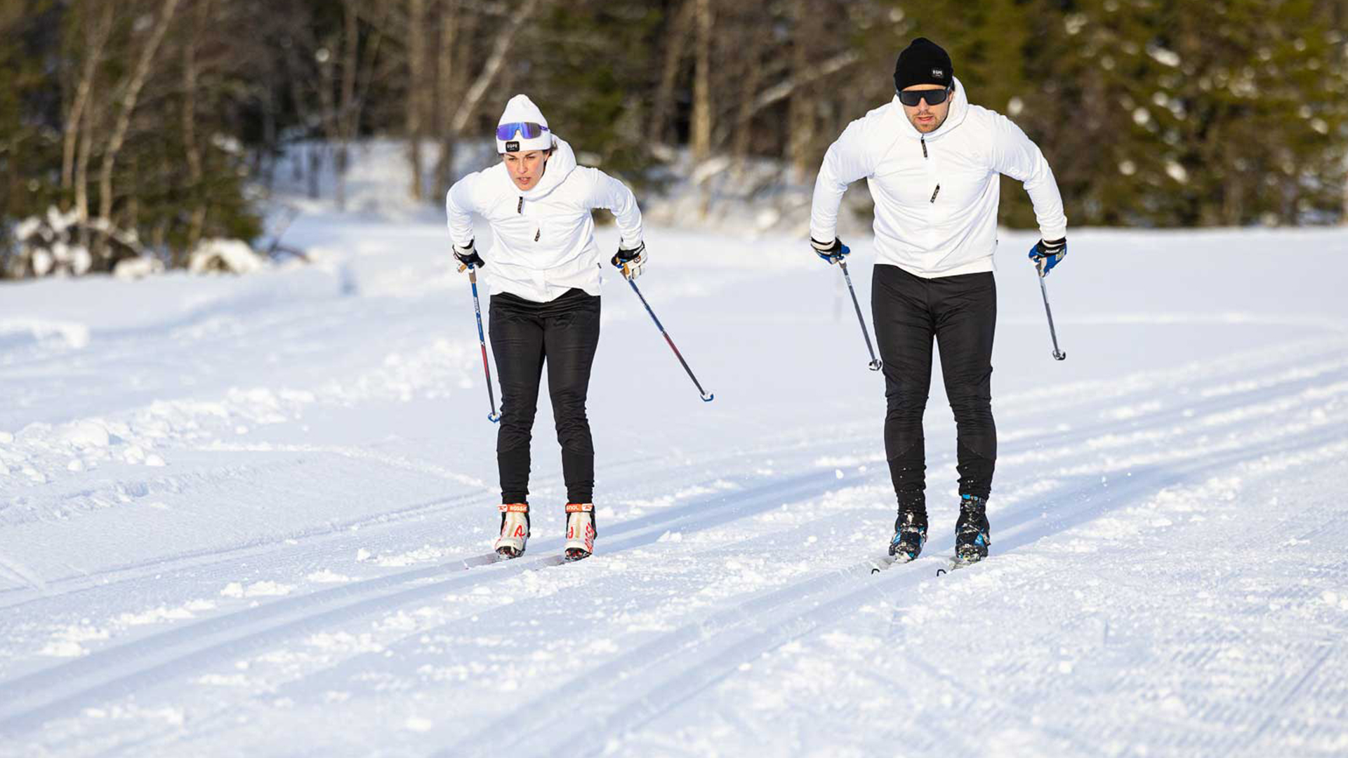Two people are cross-country skiing on a snow-covered track in a forest environment, wearing white sweaters and black pants on a sunny winter day
