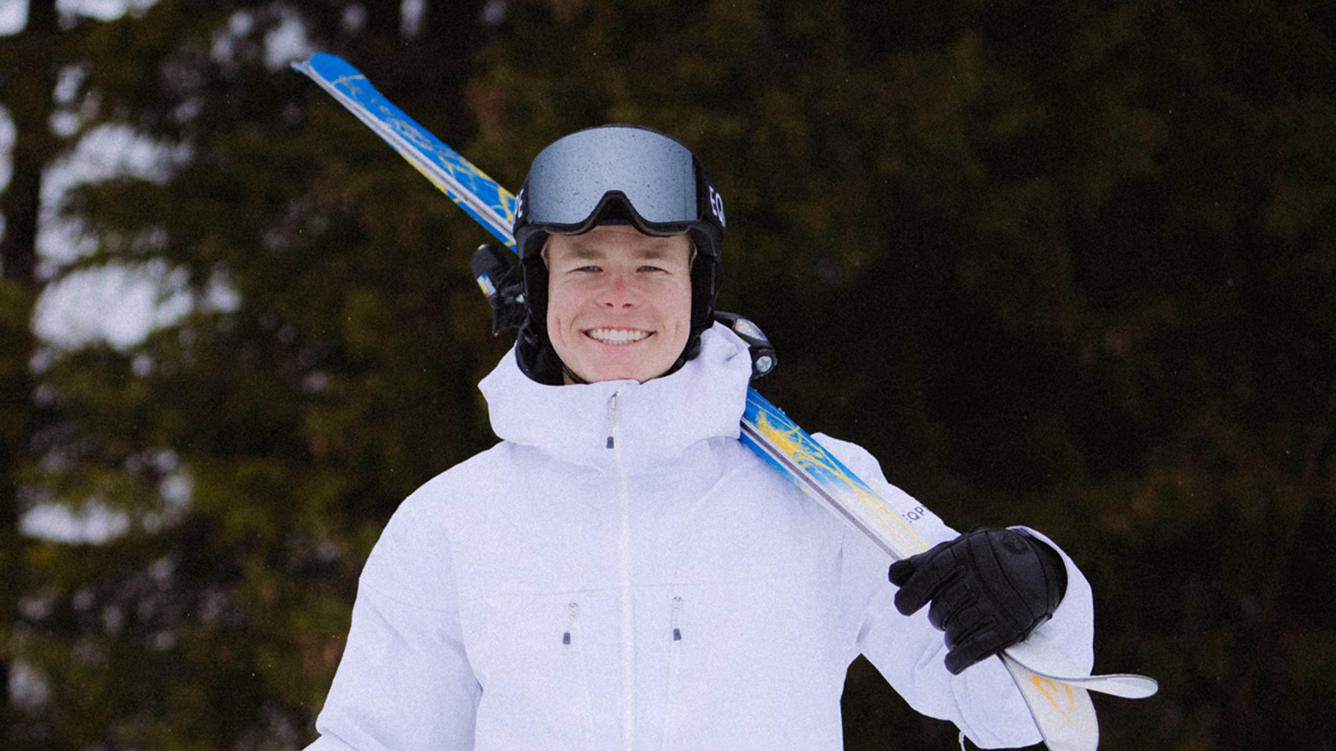 A person dressed in a white winter jacket and ski gloves, carrying a snowboard on their shoulder, stands in a forest environment