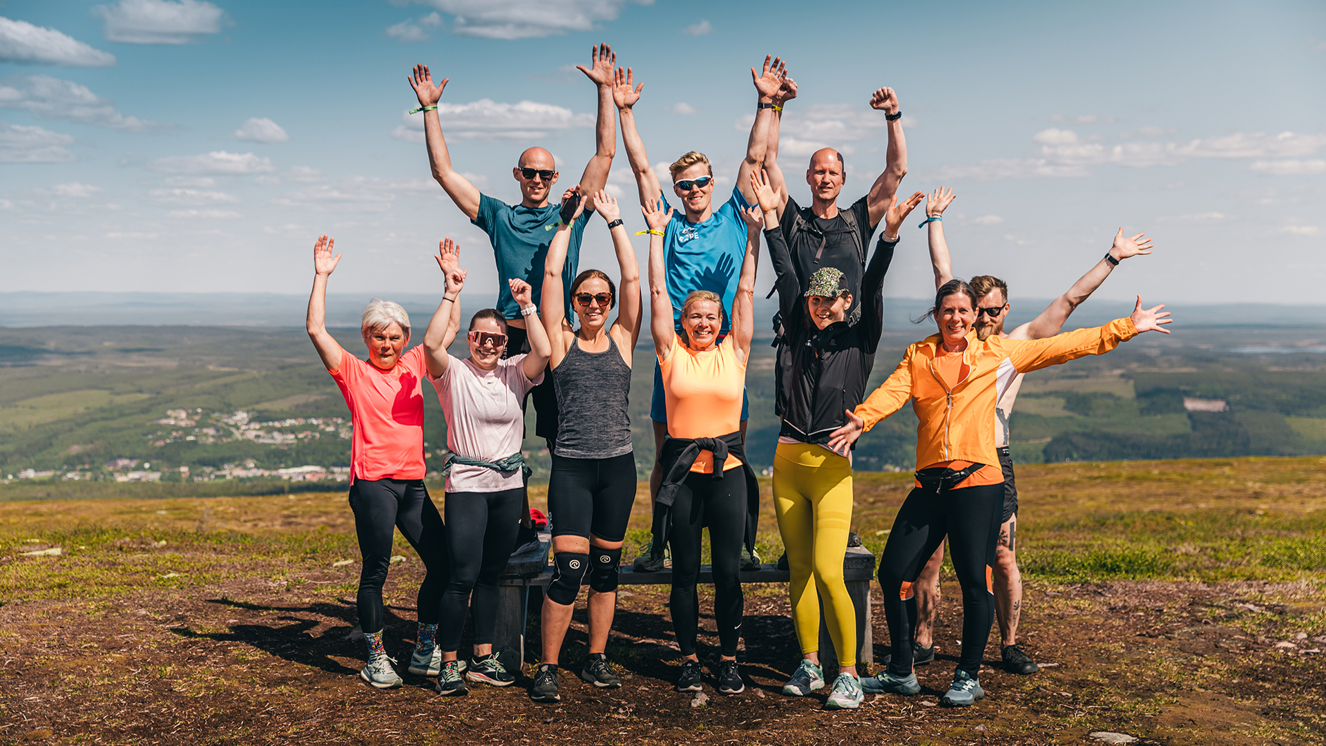 Grupp människor som sträcker upp armarna på en naturskön utsiktsplats, klädda i träningskläder under en solig dag