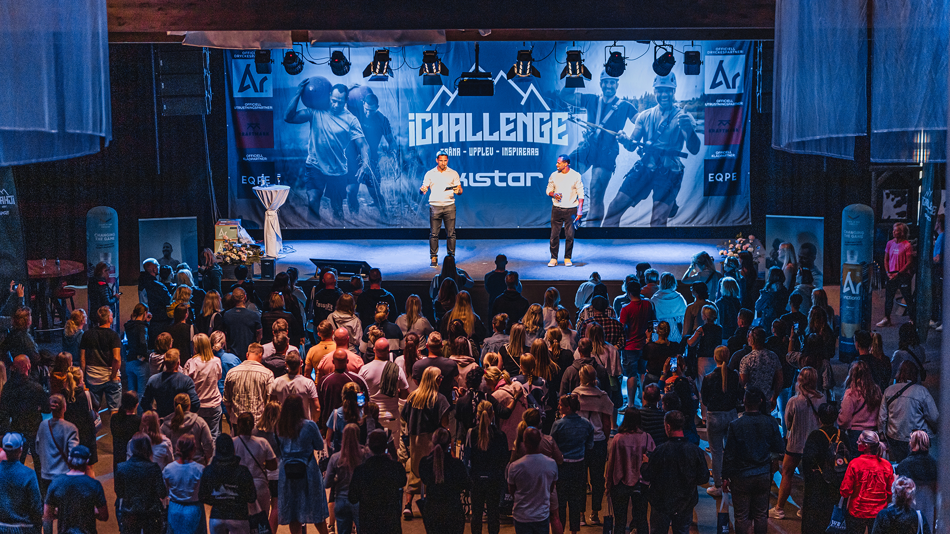 Publik på en föreläsning med "iChallenge" tema, två talare på en scen framför en stor samling åhörare i en inomhuslokal