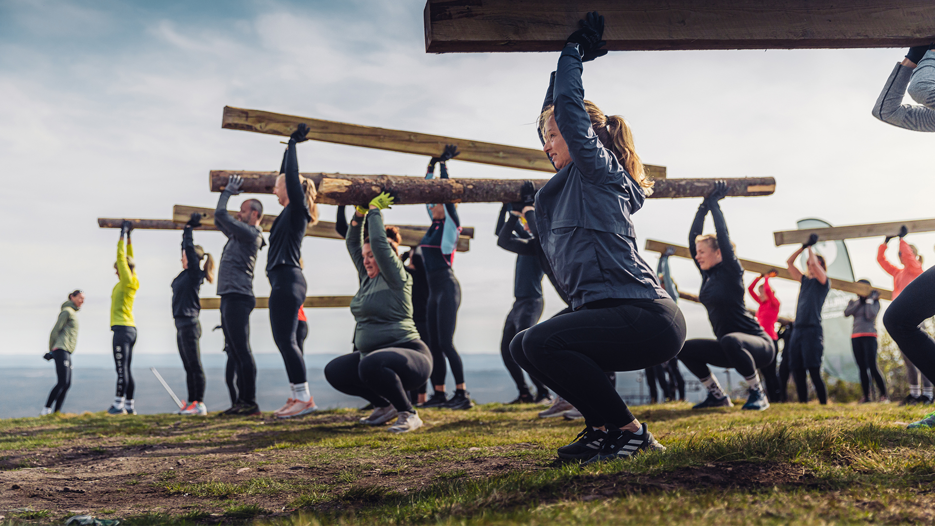 Deltagare i ett utomhusträningspass lyfter stockar tillsammans i en gruppövning på en gräsbevuxen kulle under en mulen himmel