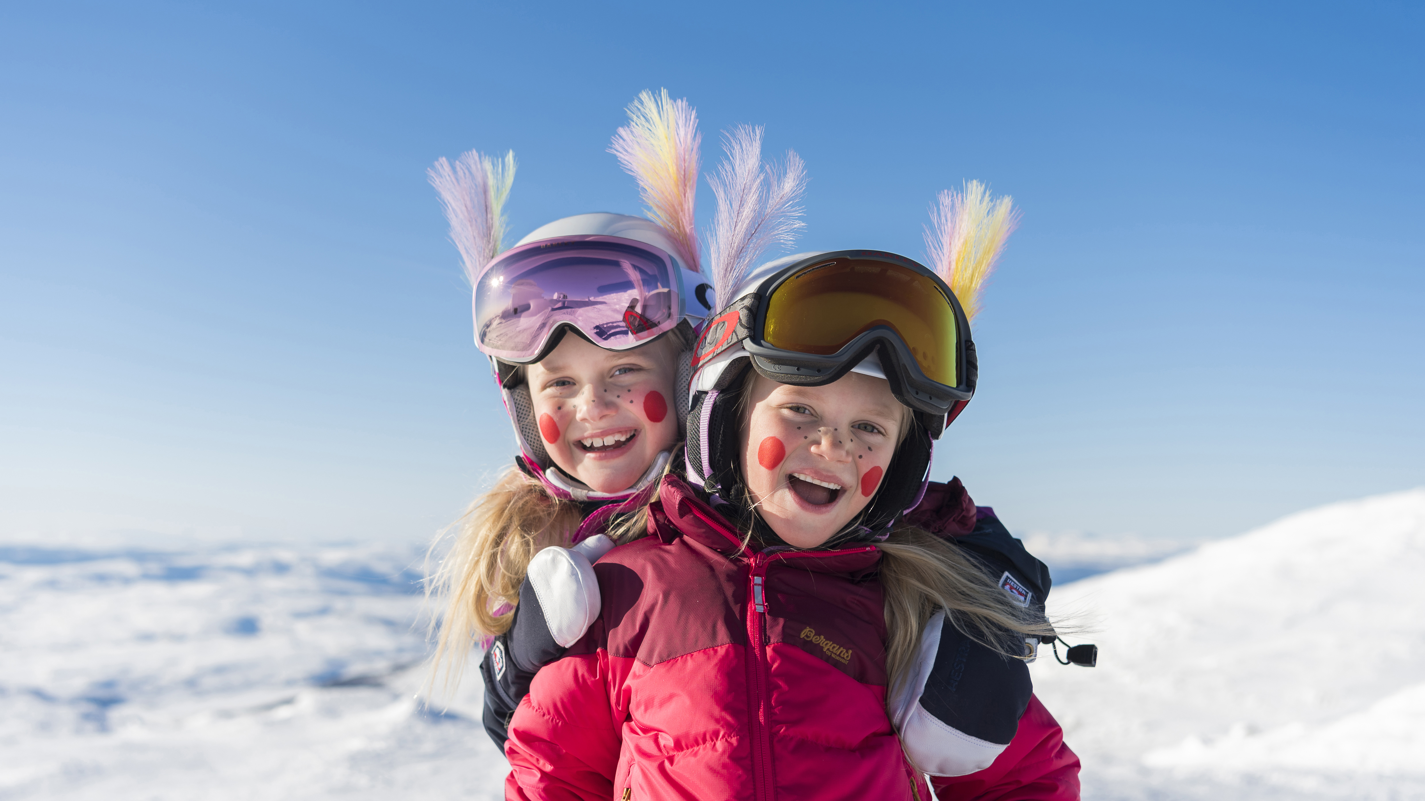 Två leende barn med färgglada hjälmar och skidglasögon står i snön mot en klarblå himmel. Påskfjädrar på hjälmarna.