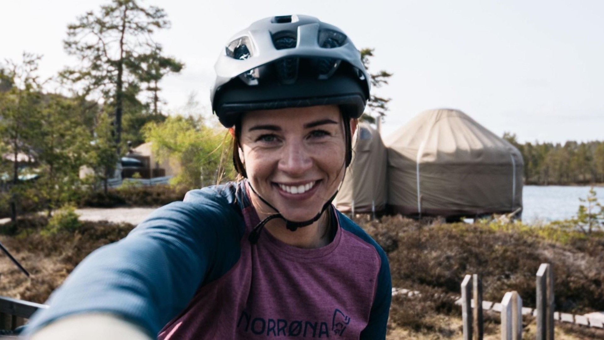 Kvinde i cykelhjelm smiler udenfor nær telt og sø