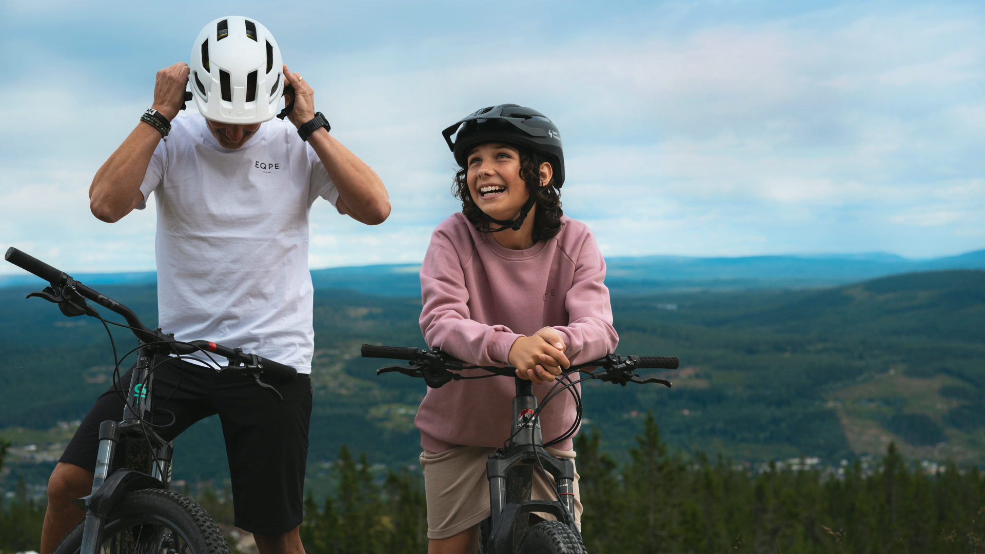 Två personer på mountainbikes njuter av en cykeltur i en naturskön bergsmiljö. Den ena justerar sin hjälm medan den andra ler mot kameran