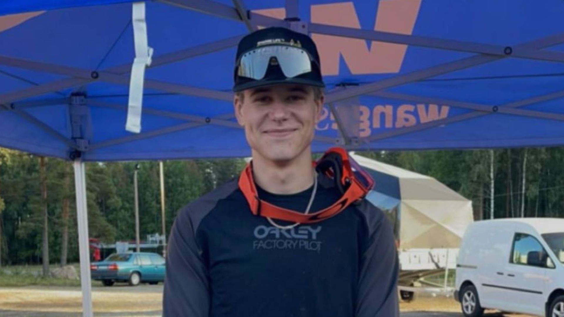 Person i sort trøje og solbriller står under en blå teltdug udendørs. Baggrunden viser træer og en parkeret hvid varevogn