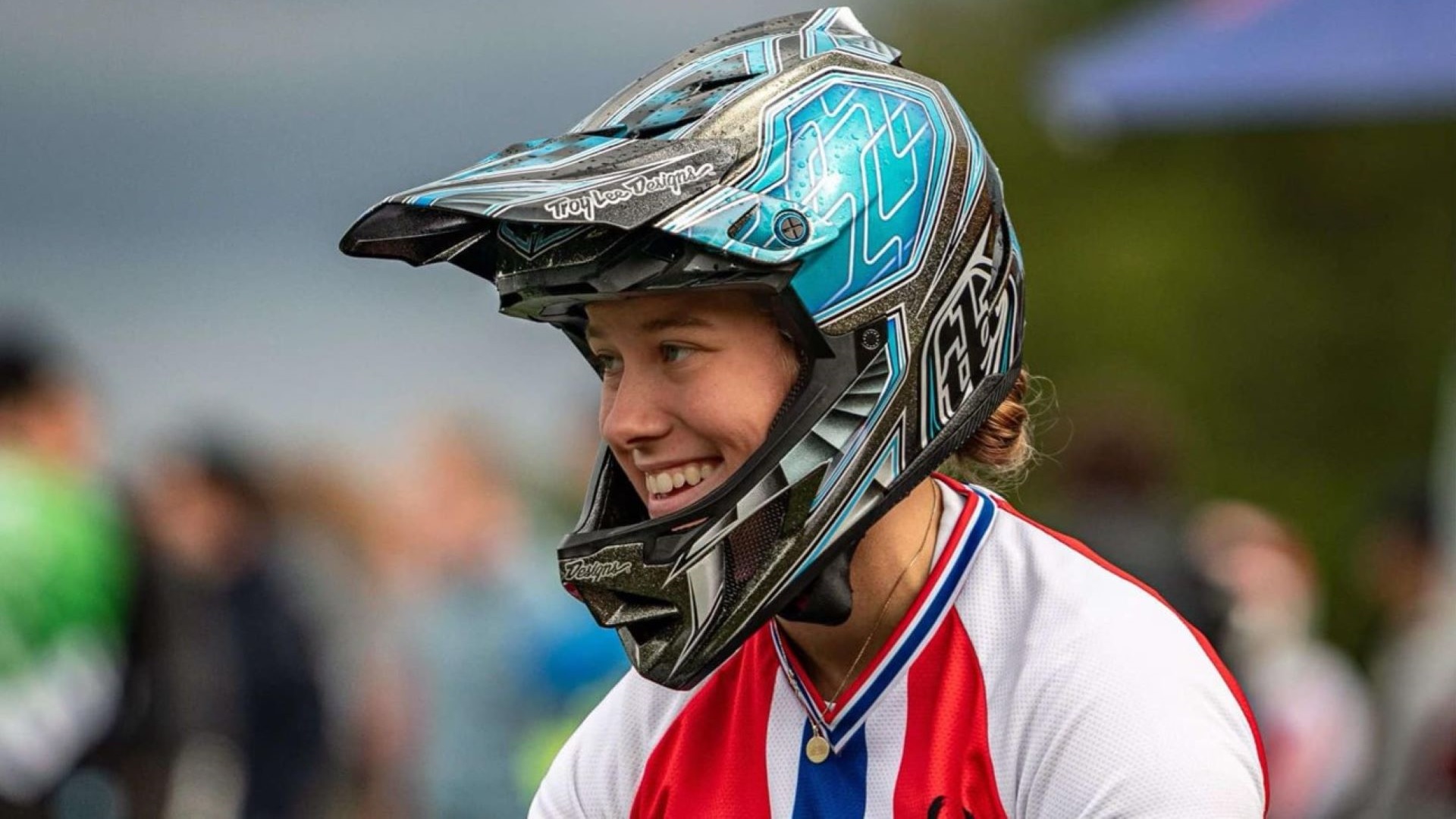 Kvinde i BMX-hjelm smilende iført rød og hvid trøje ved sportsbegivenhed