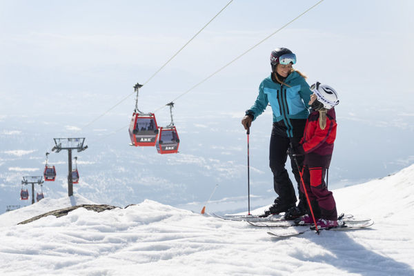 To skiløbere står på en sneklædt bjergskråning med røde svævebaner i baggrunden ved et skisportssted