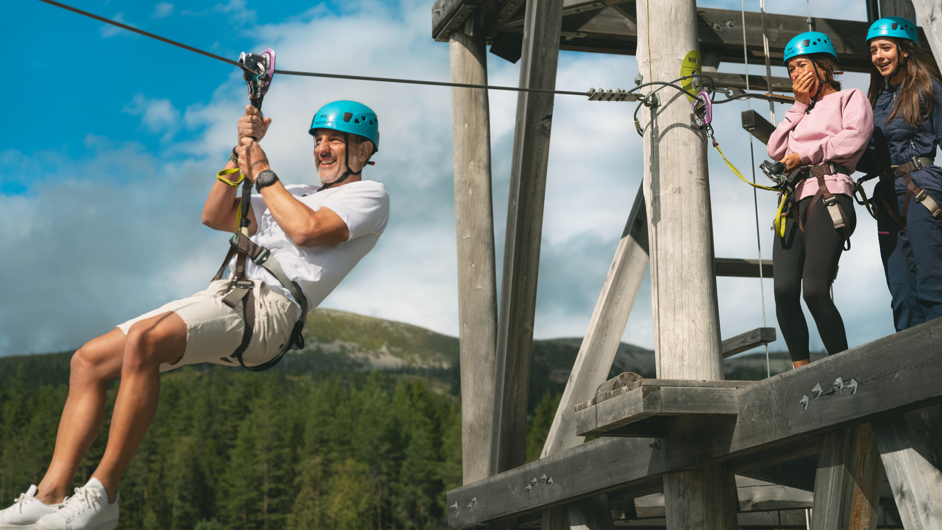 Man åker linbana med säkerhetsutrustning medan två personer tittar på från plattformen