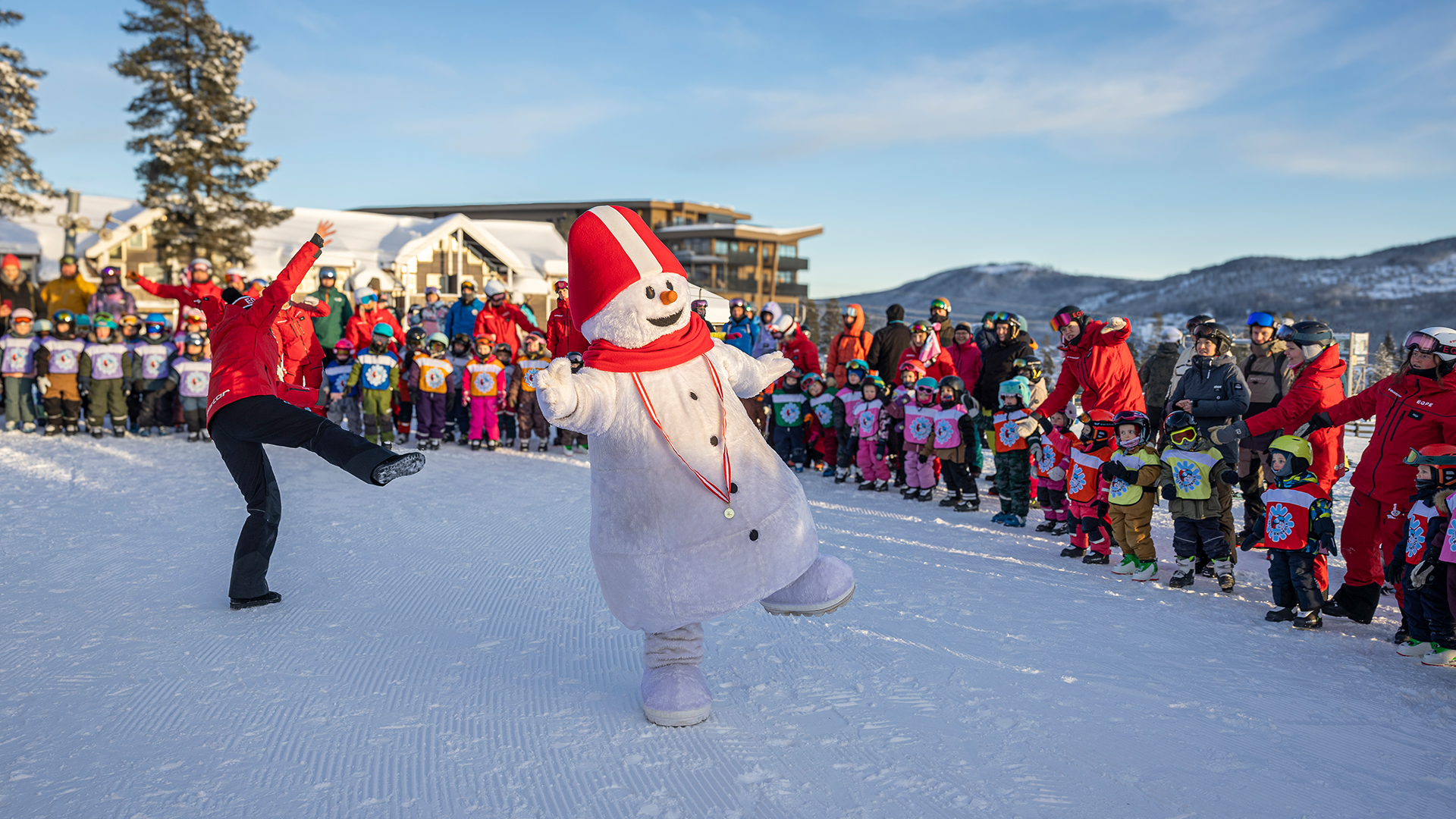 Snögubbe dansar med barn och instruktörer på en skidort, omgiven av snö och soligt väder