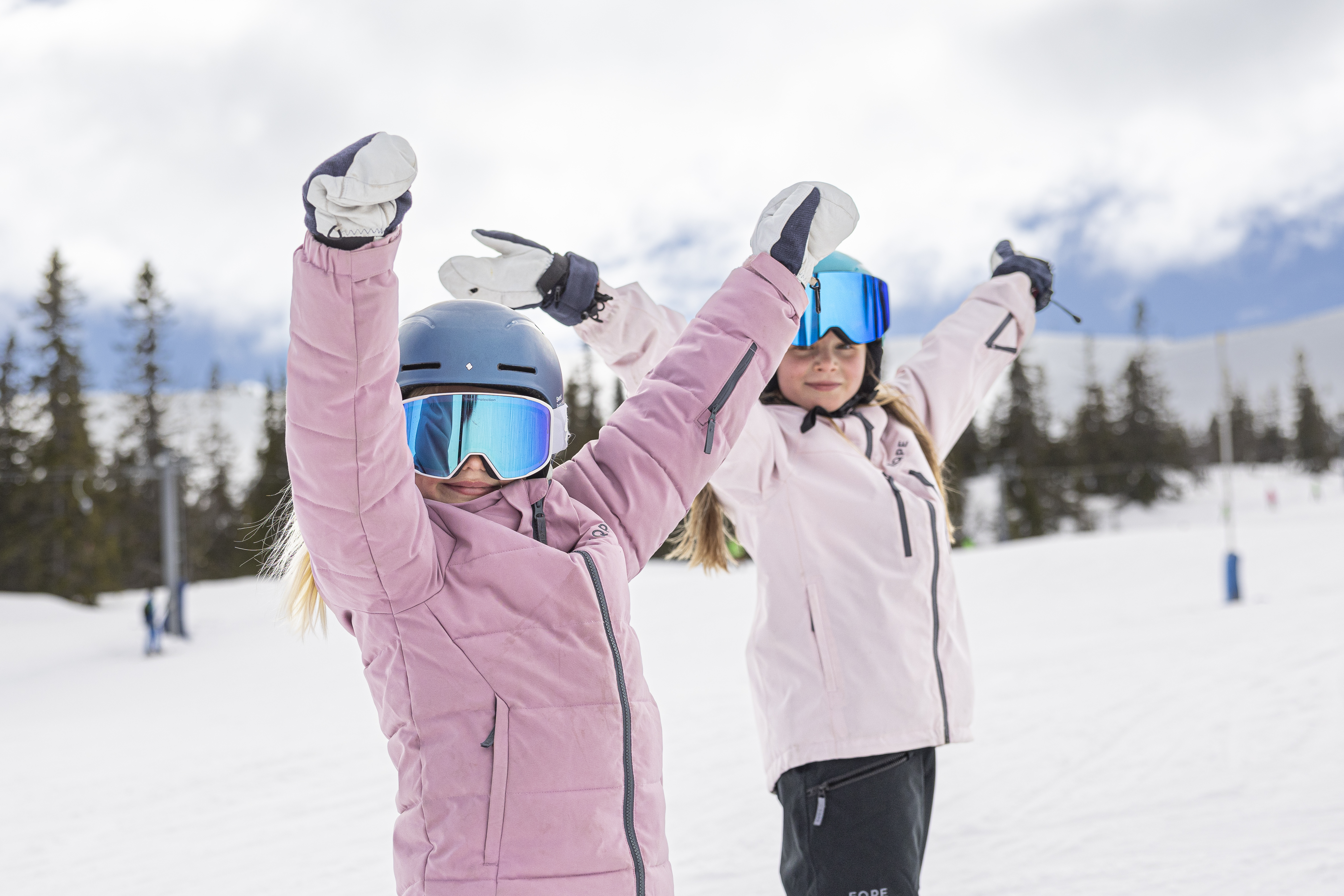 Två barn i rosa vinterkläder och skidglasögon åker skidor i en snötäckt bergsmiljö