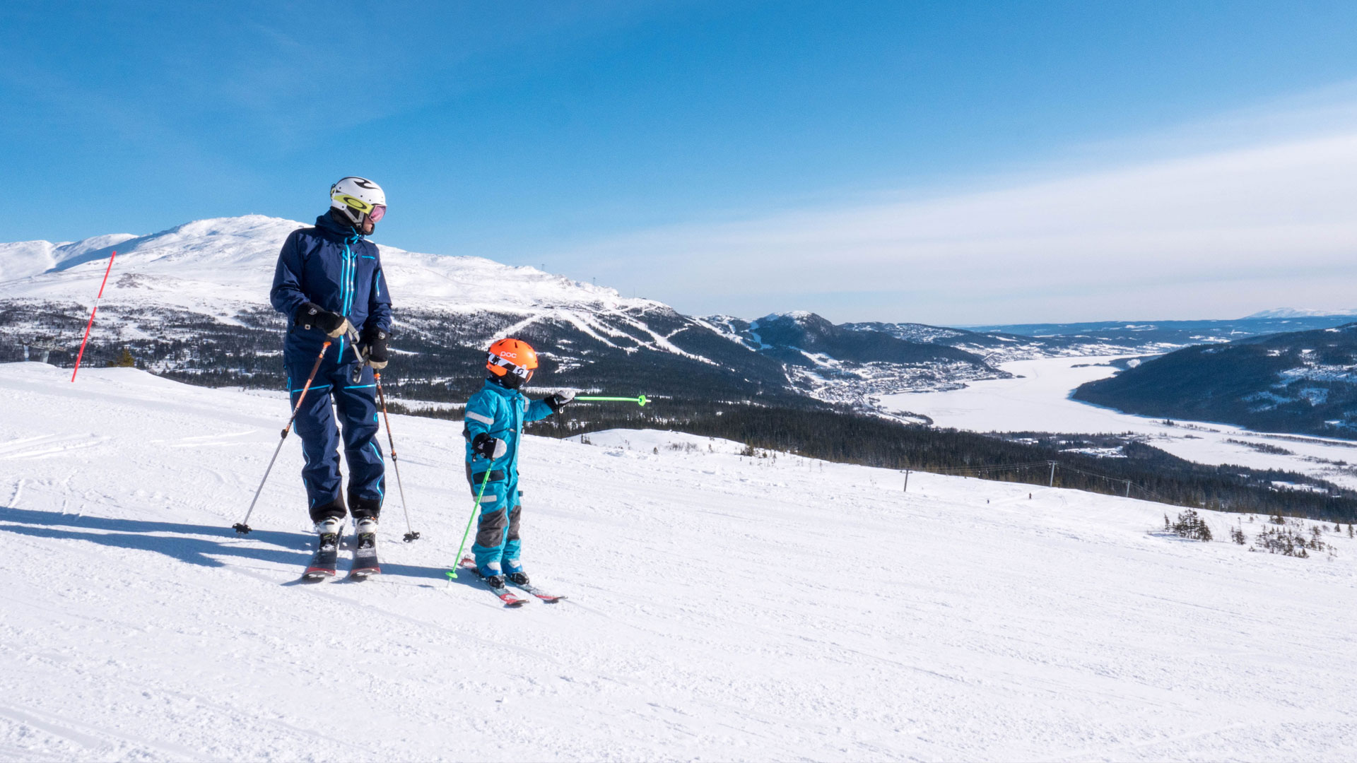 Två skidåkare på en solig skidbacke i fjällen, med en vidsträckt utsikt över snötäckta berg och dalar i bakgrunden