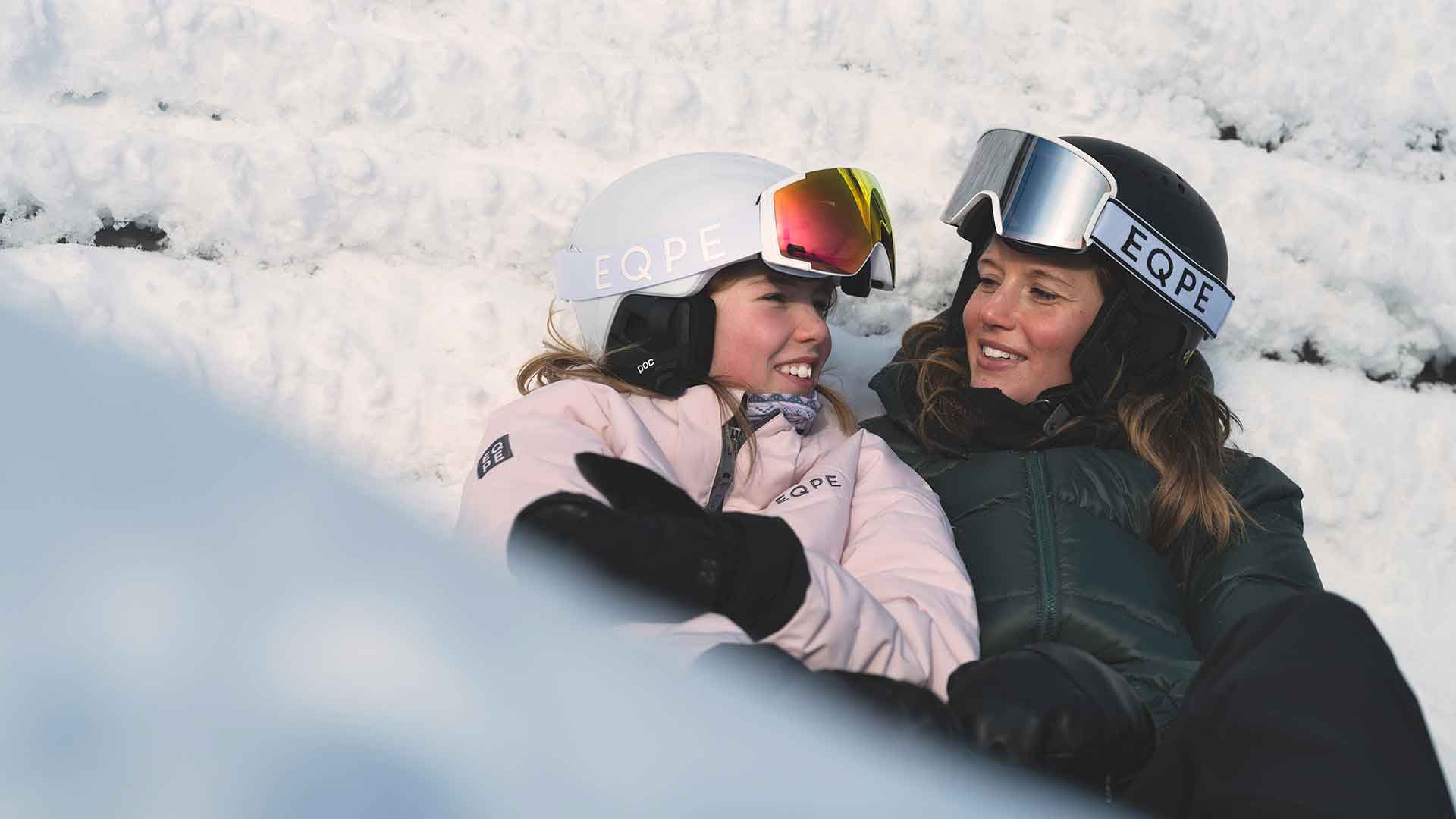 To personer i vintertøj og hjelme med EQPE-logo, sidder og nyder i sneen