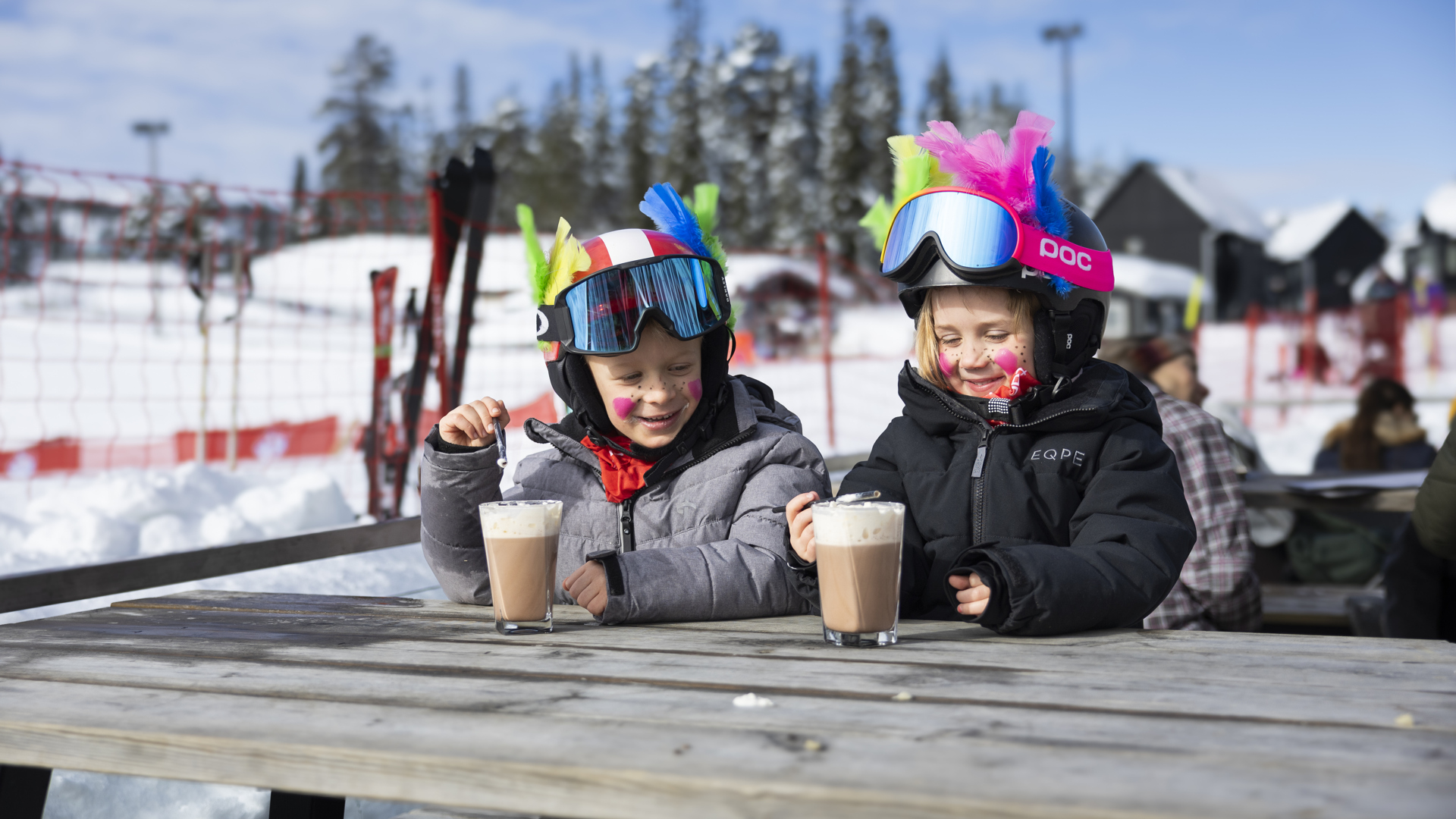Två barn på påsklov i fjällen njuter av varm choklad vid ett utomhusbord, klädda i skidkläder och pyntade med fjädrar.