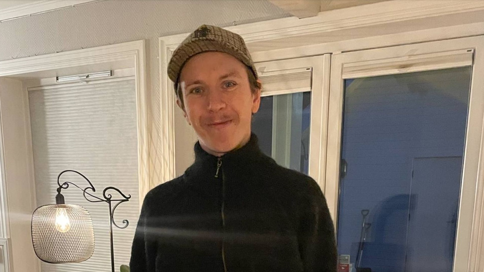 En person i sort trøje og kasket står indendørs ved siden af en lampe med netmønster. Baggrunden viser store vinduer