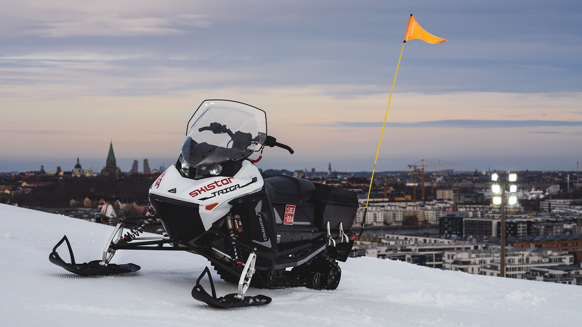 Snöskoter SkiStar e-TraX parkerad på en snötäckt kulle med en stad i bakgrunden, utrustad med en orange säkerhetsflagga