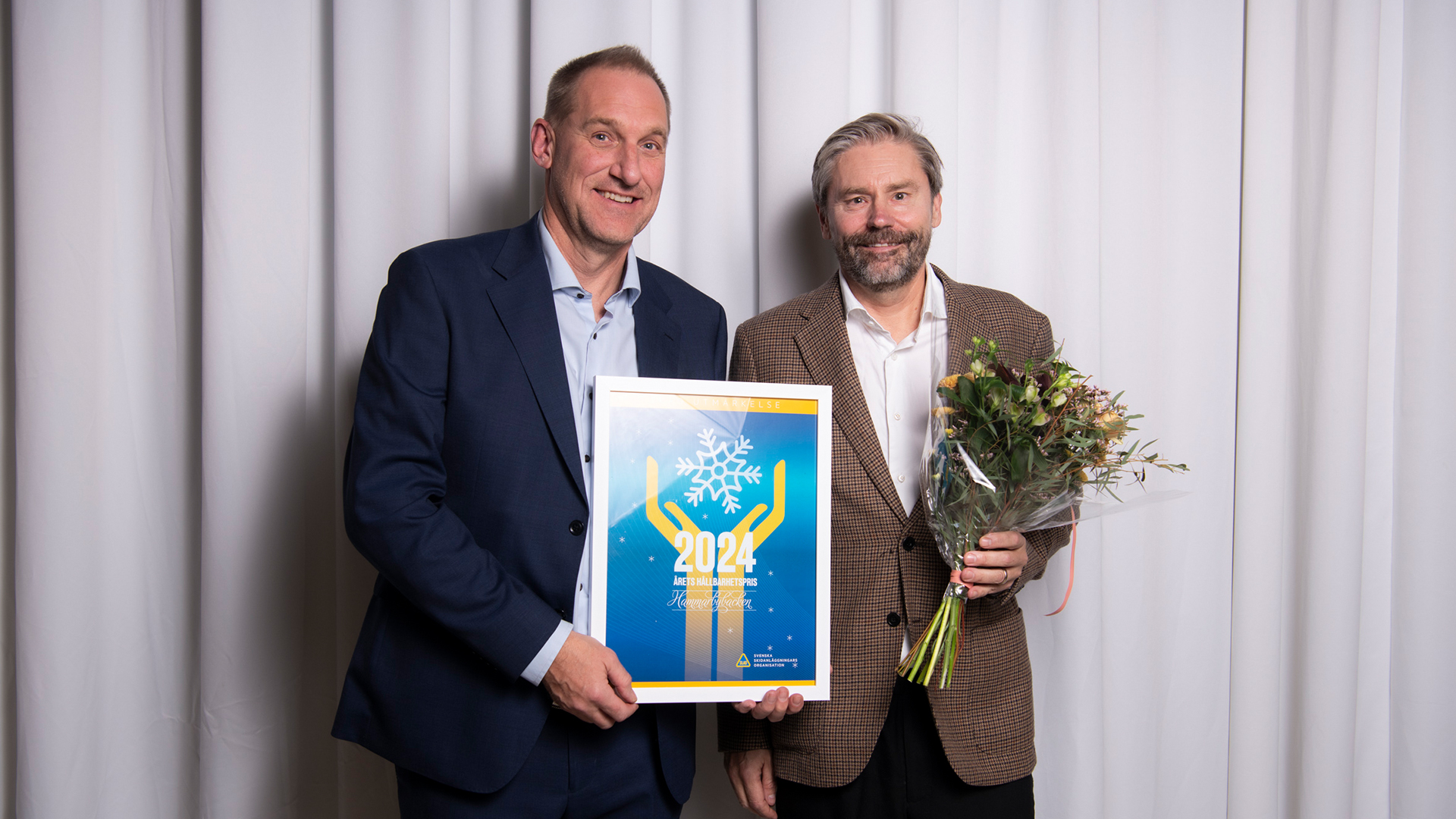 Två män poserar glatt framför en vit bakgrund; en håller i ett diplom och den andra håller en blombukett, vilket antyder en prisutdelning eller ceremoni