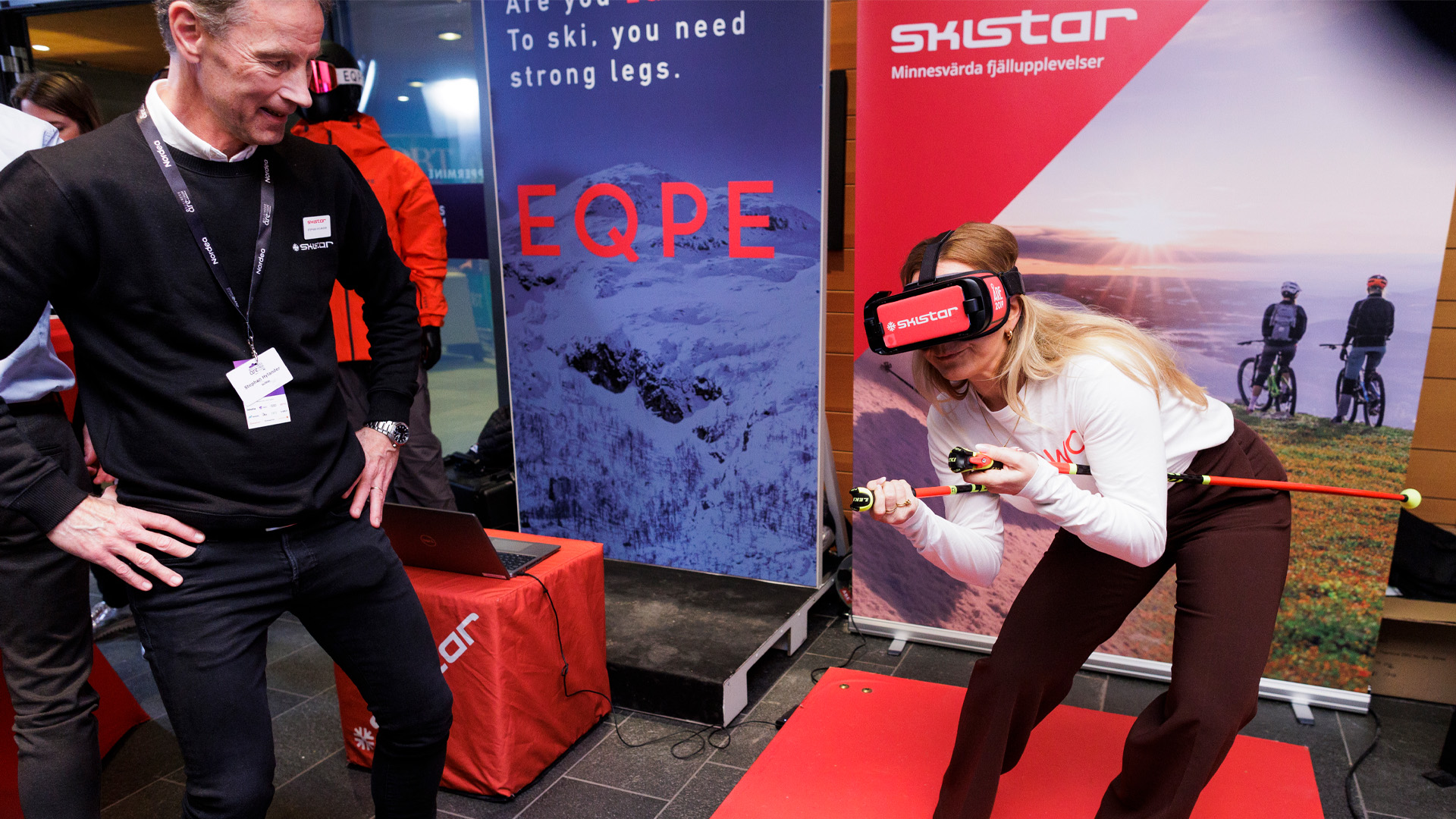Kvinna använder VR-headset för skidåkning med SkiStar-utrustning på mässa, omgiven av reklamaffischer och en manlig åskådare