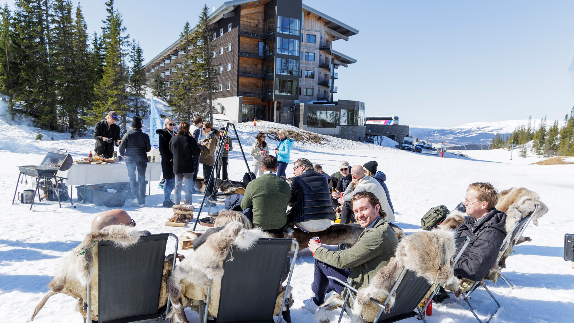 Personer sitter utomhus och njuter av soligt väder i snön framför ett modernt hotell i fjällen. De verkar socialisera vid ett grillområde med omgivande vinterlandskap